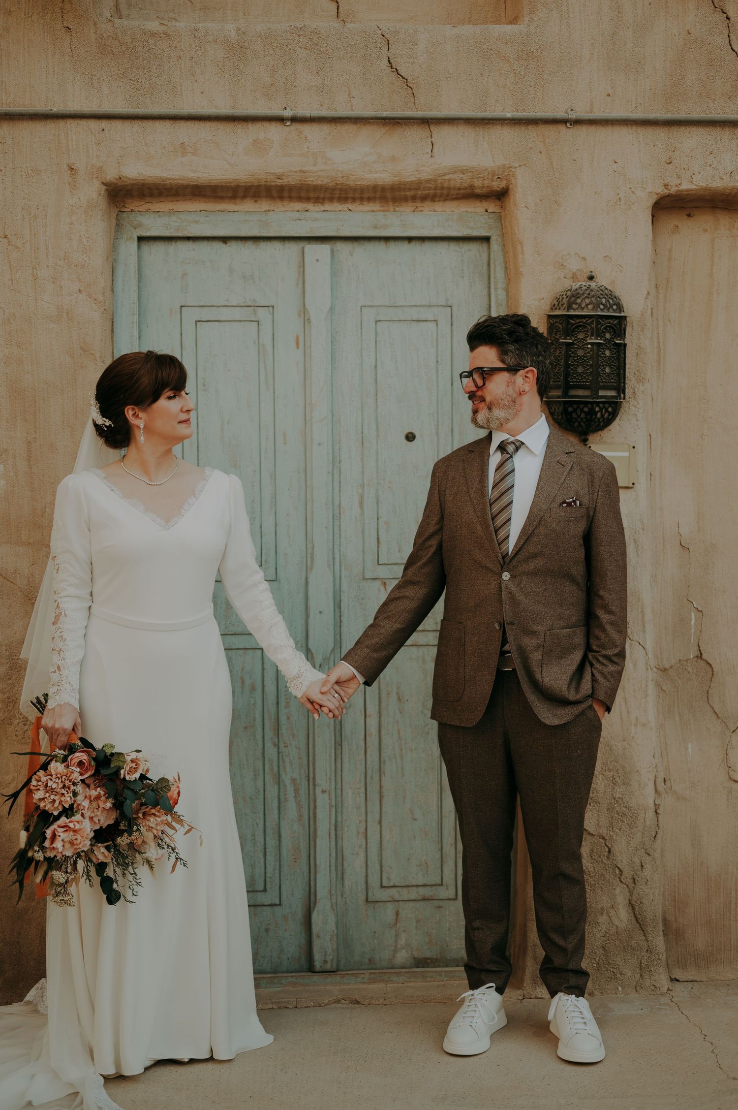 A couple in wedding attire poses against a weathered wall with vintage blue doors.