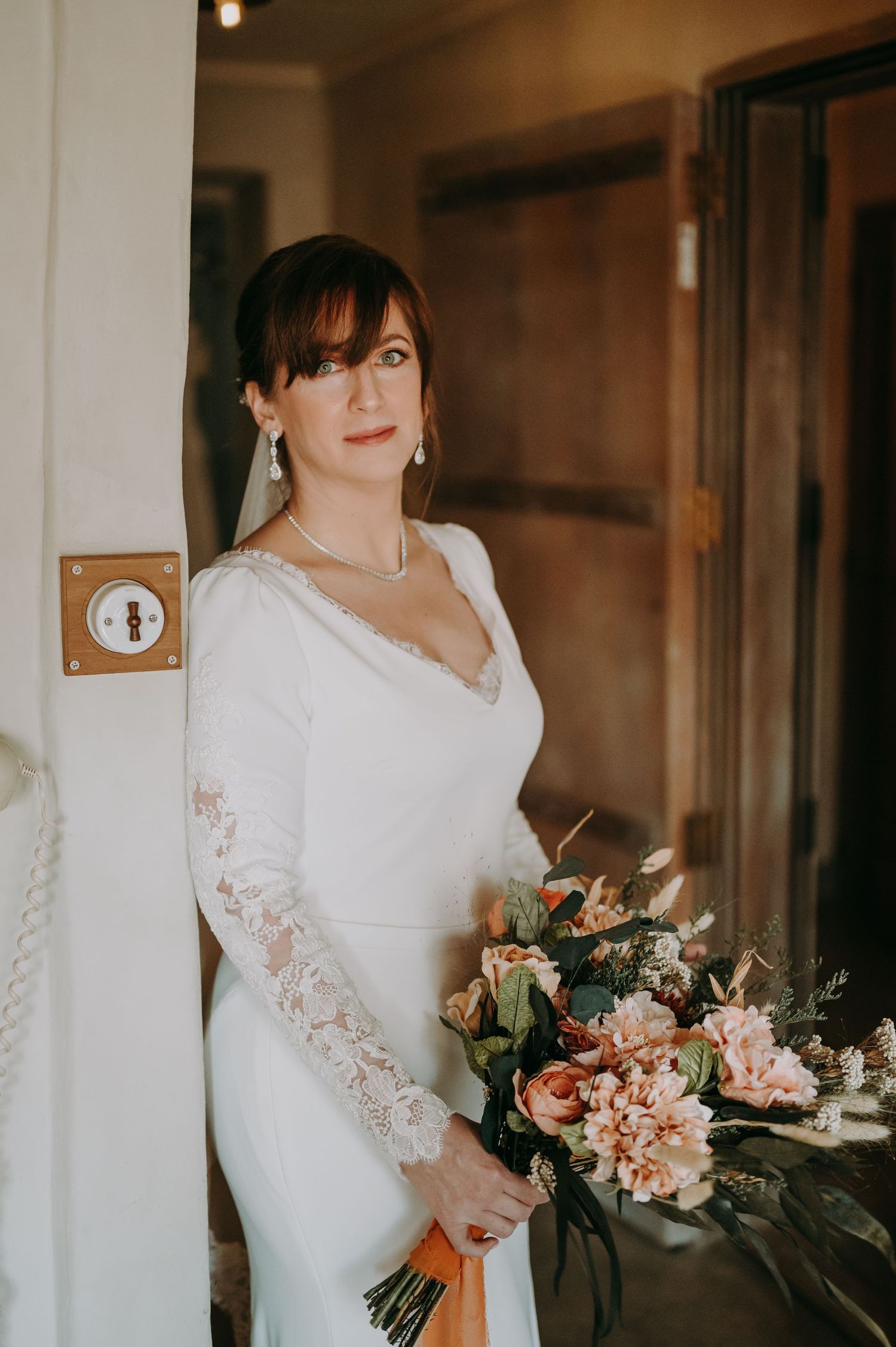 A bride in an elegant white lace gown holds a romantic bouquet of pink and peach flowers.