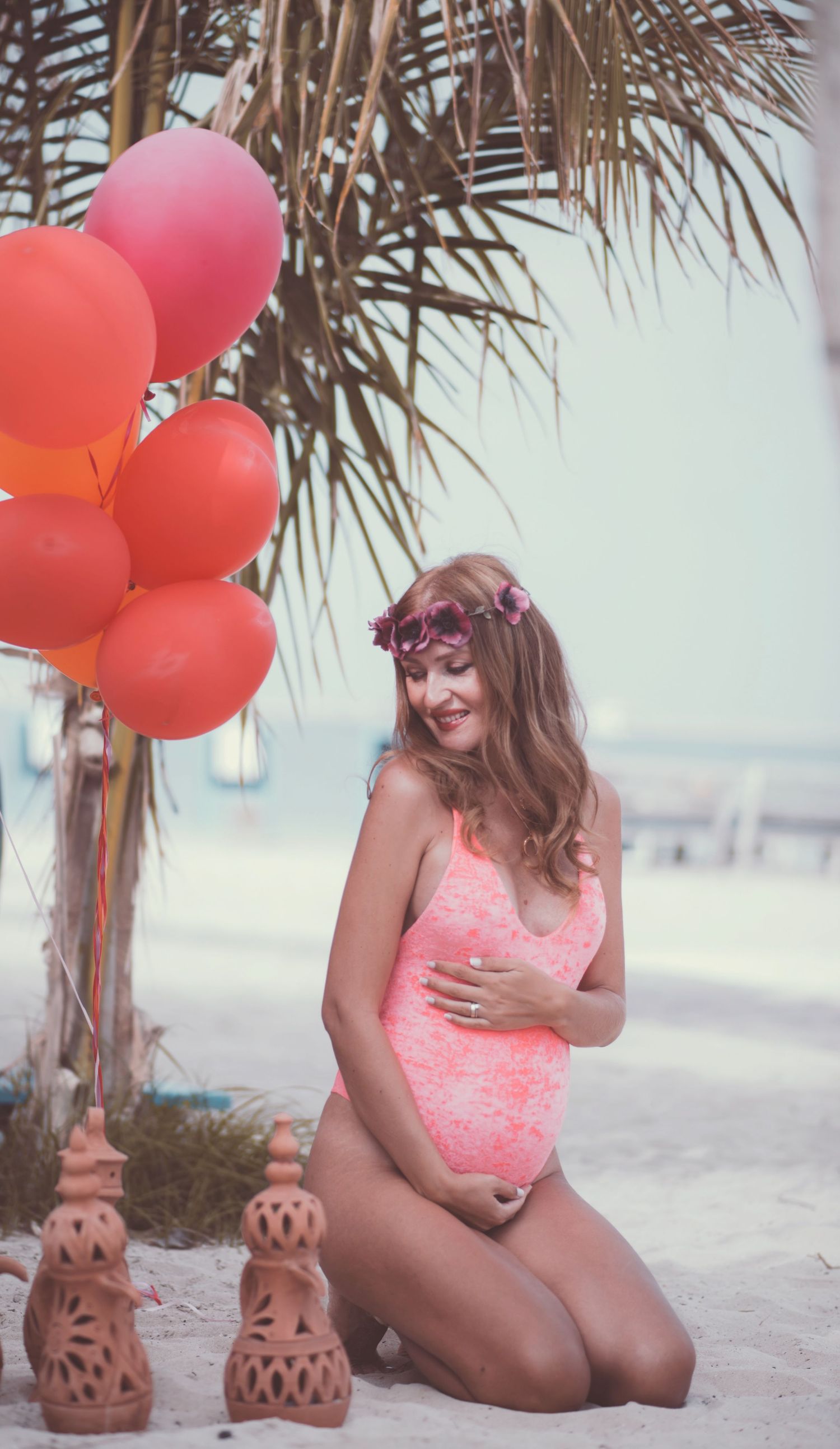 Beach maternity photoshoot with red balloons and palm trees in soft lighting.