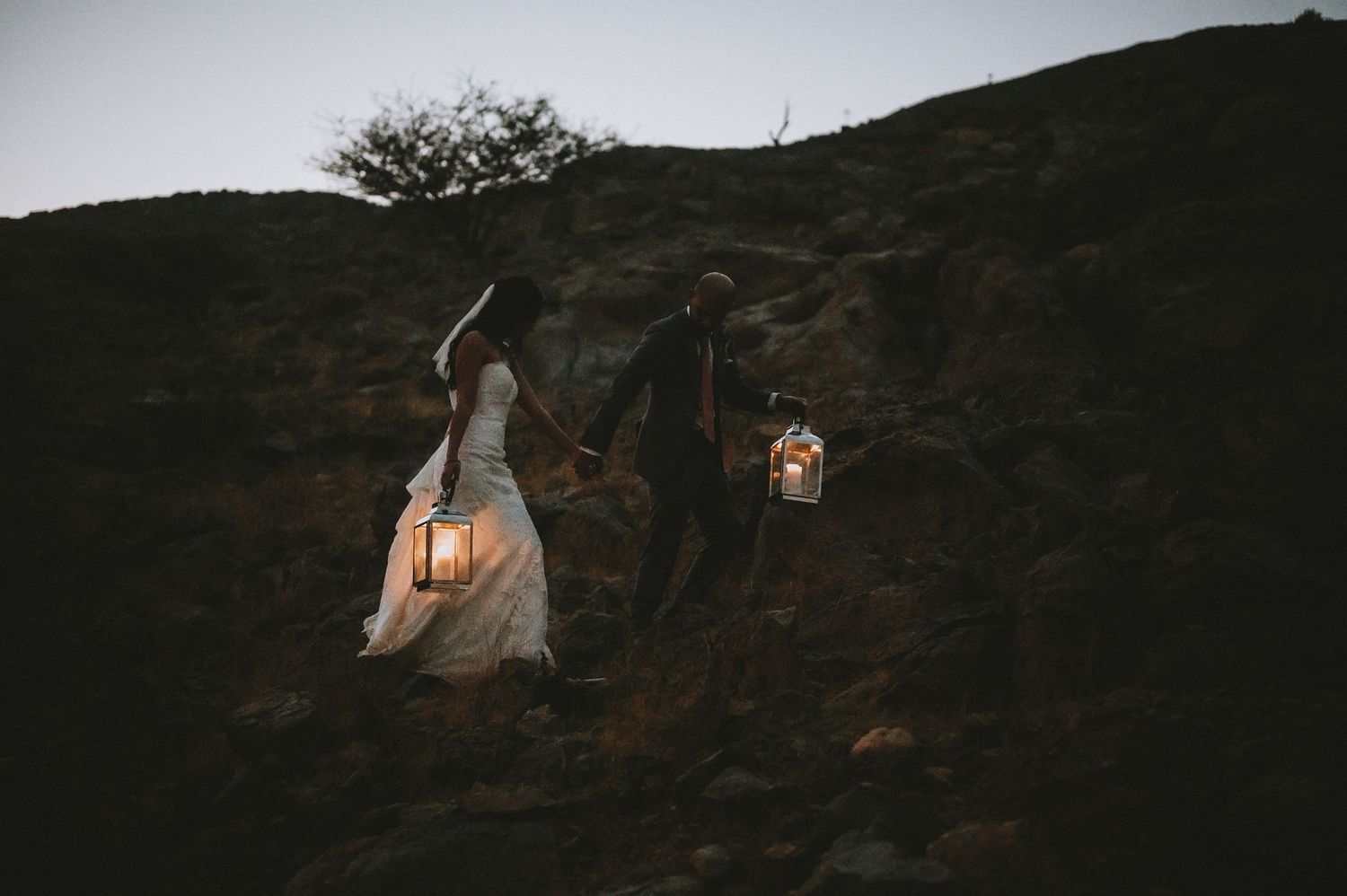 Wedding couple stands on rocky terrain at dusk holding vintage lanterns against a moody sky.