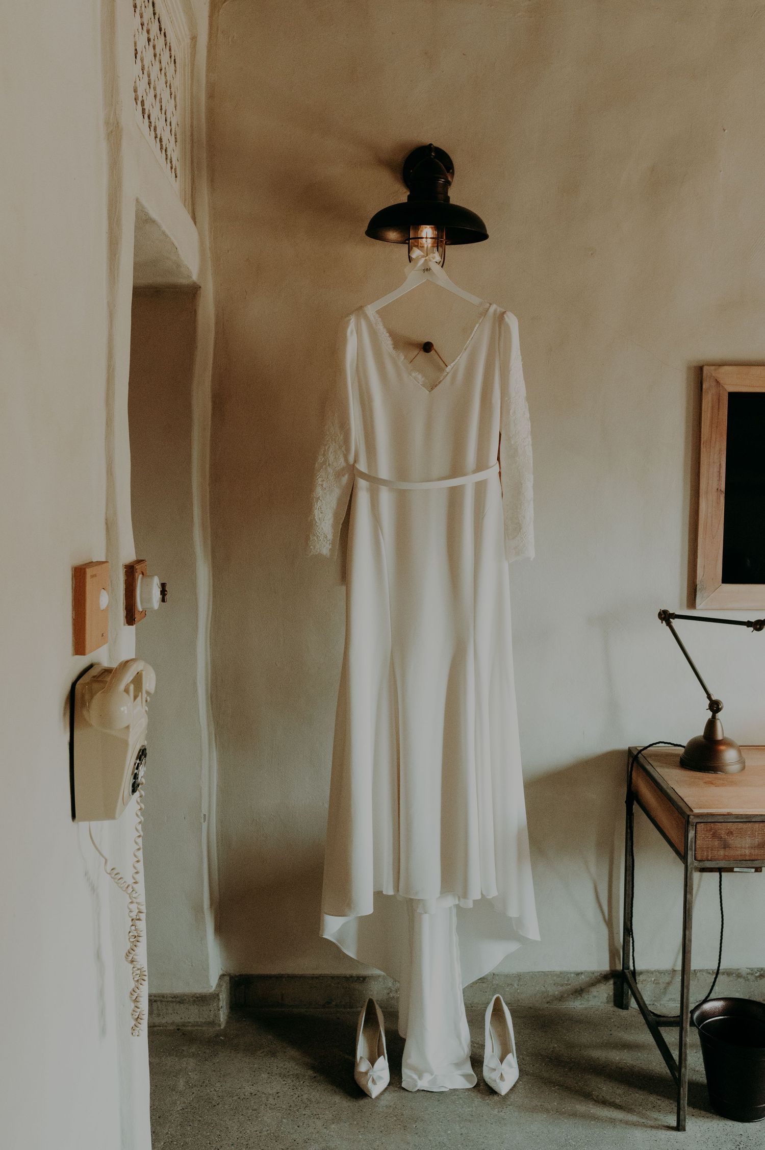 An elegant white wedding dress hangs displayed against a neutral wall in soft natural light.