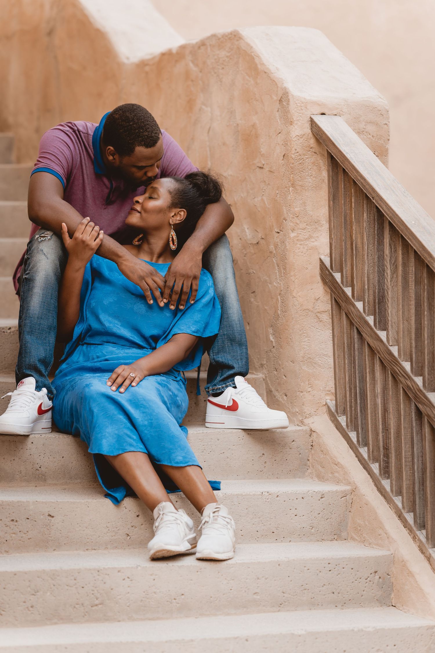 A couple embraces intimately while sitting on concrete stairs in casual attire.