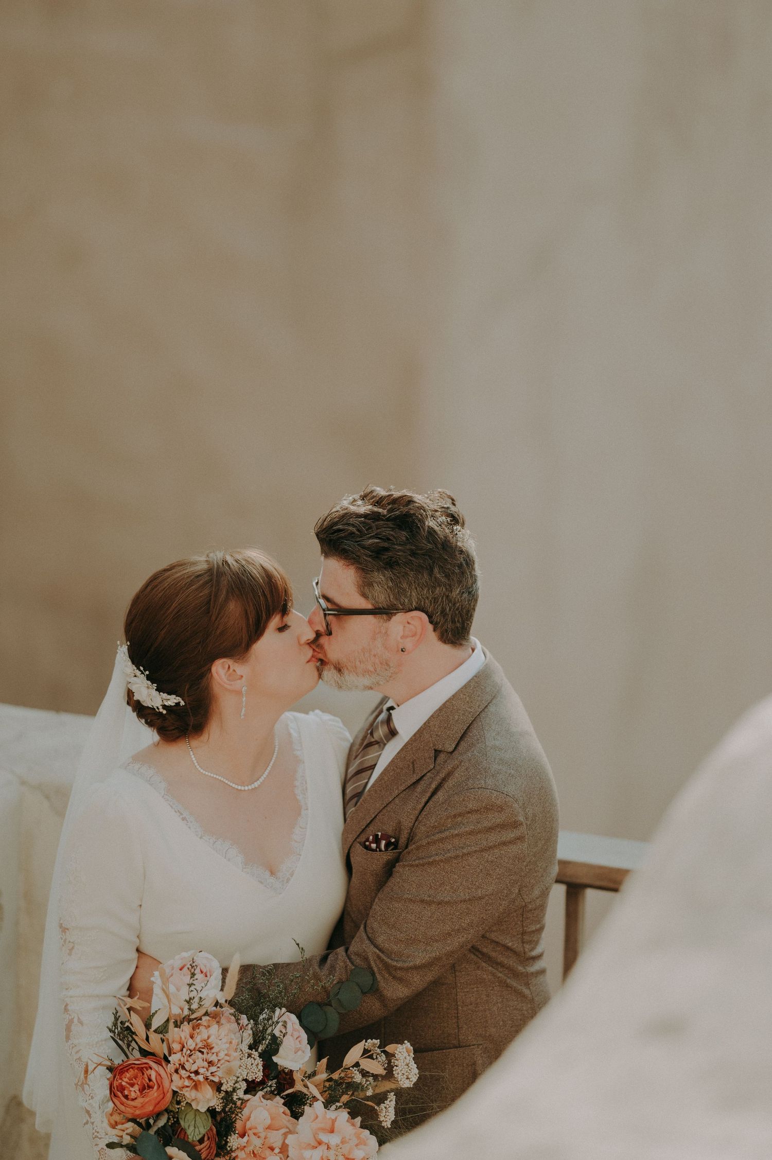 Romantic wedding couple sharing an intimate moment with coral bouquet against neutral background.