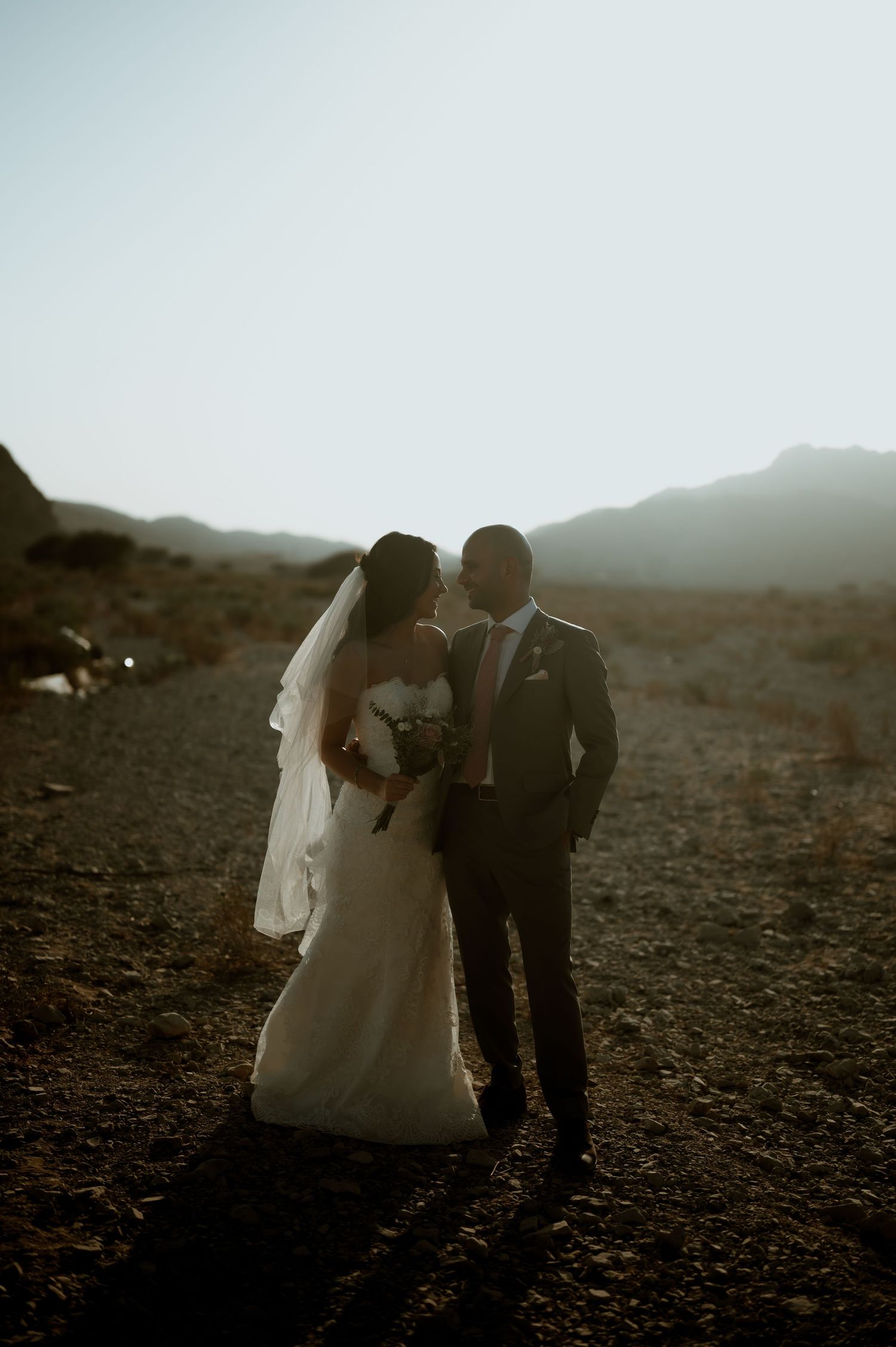 Couple shares intimate moment in desert with dramatic mountain silhouette behind them.