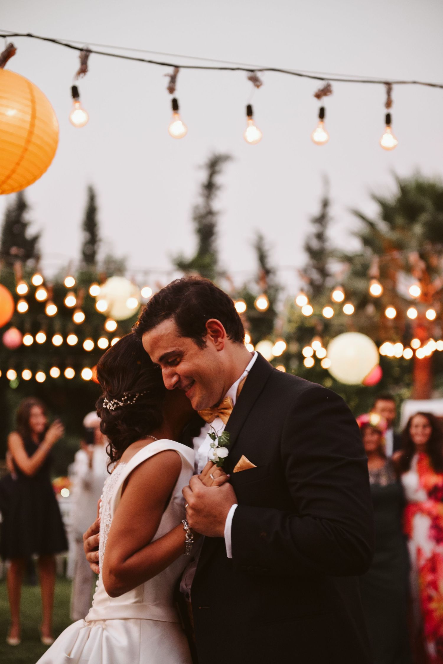 Romantic evening dance under string lights and paper lanterns at an outdoor reception.