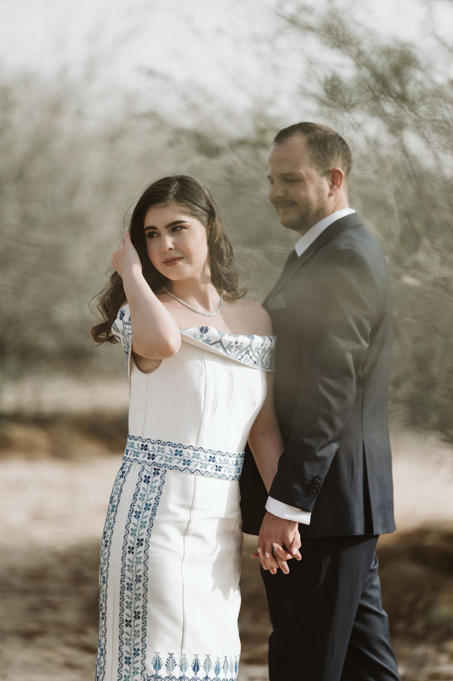 A wedding couple poses together outdoors, with the bride wearing a distinctive white dress with blue details.