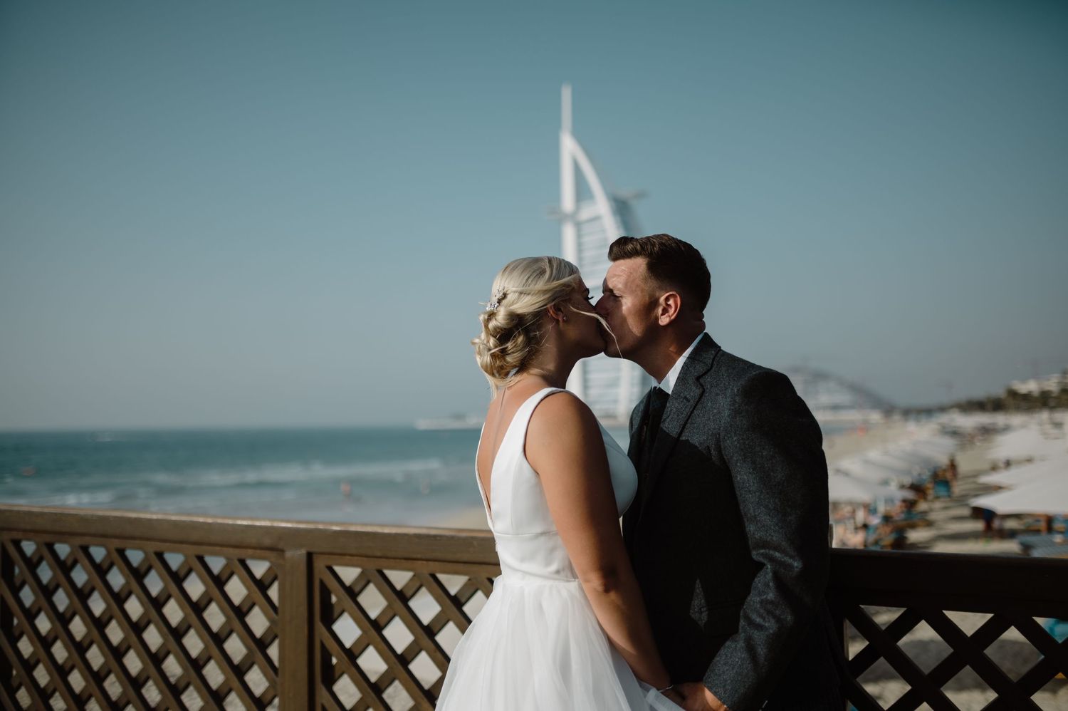 Couple embracing on a beachfront boardwalk with palm trees and ocean views at sunset.