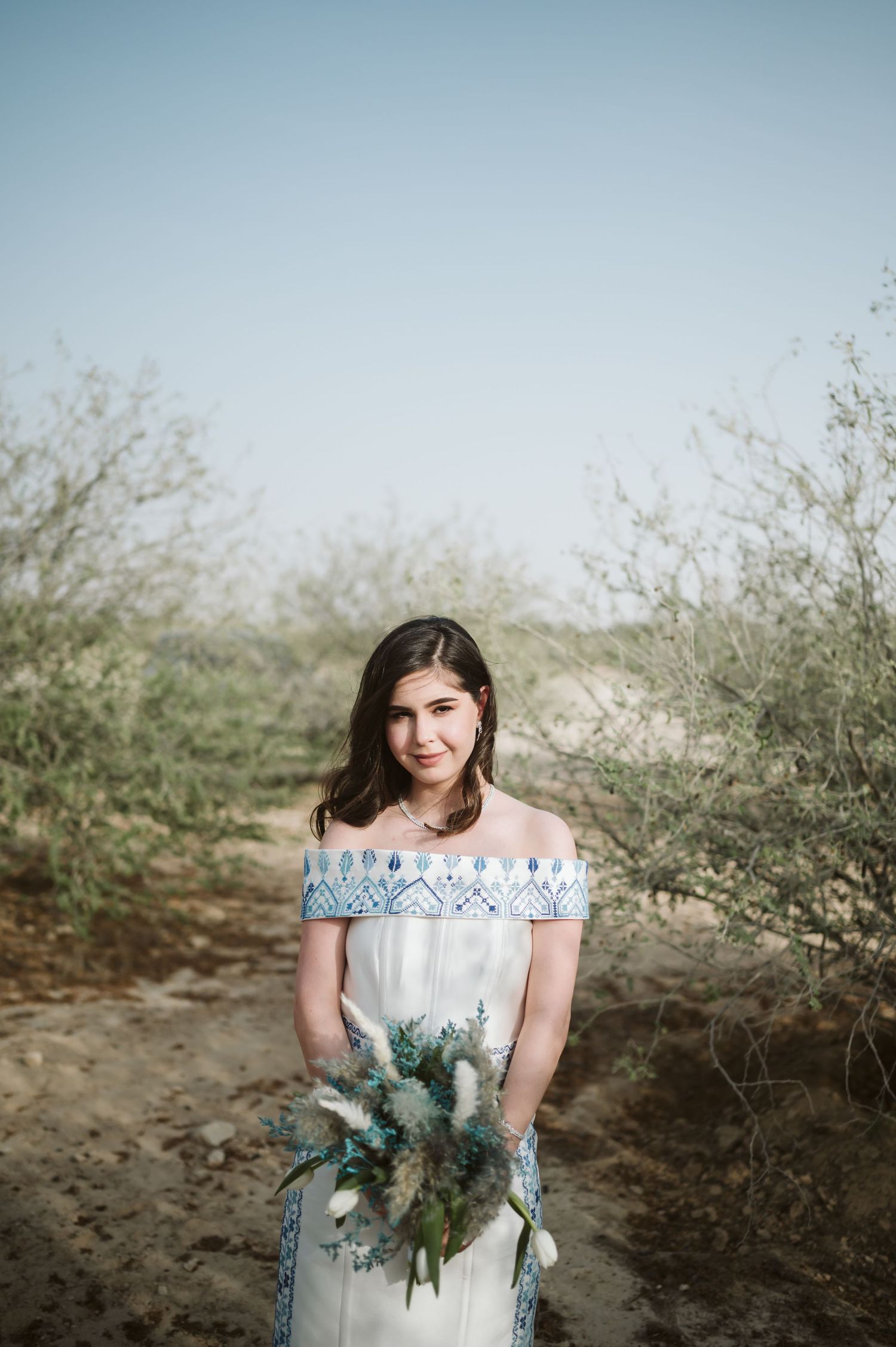 Woman in white lace dress holds bouquet in desert setting with brush background.
