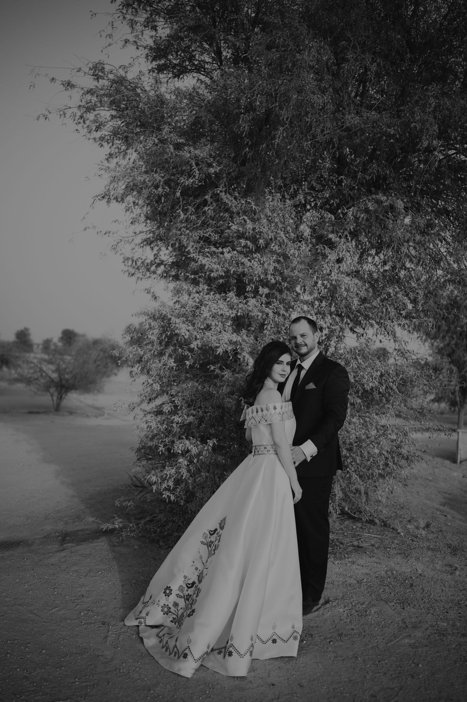Black and white wedding photography captures a couple's romantic moment under trees in a desert setting.