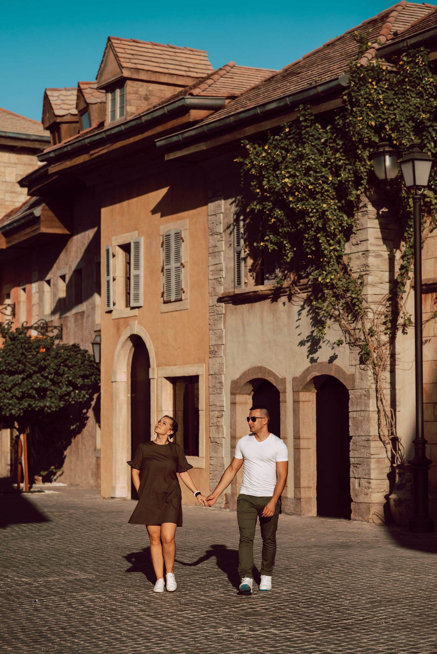 Two people walk through a charming European cobblestone street lined with historic stone buildings and archways.