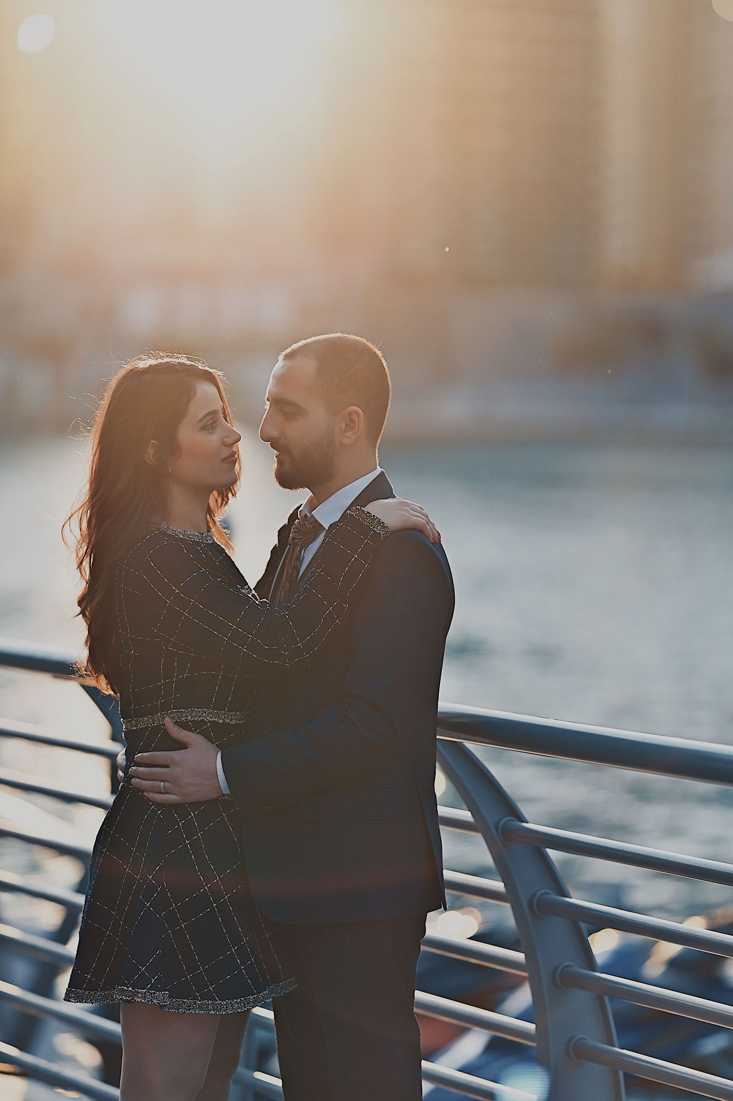 Couple embracing on waterfront with soft golden light and urban backdrop.