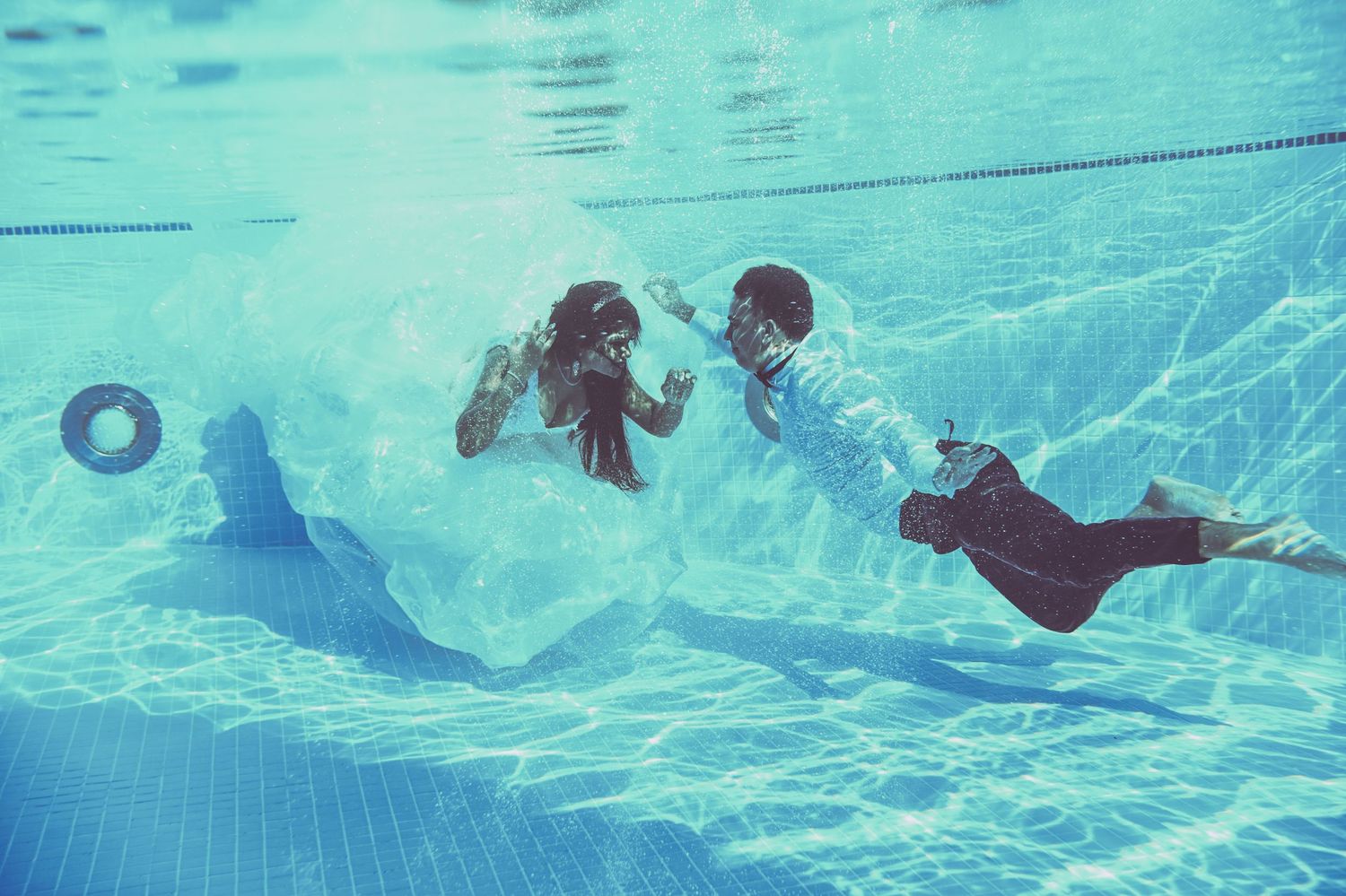 Couple in formal attire swimming underwater in a pool with ethereal blue lighting.