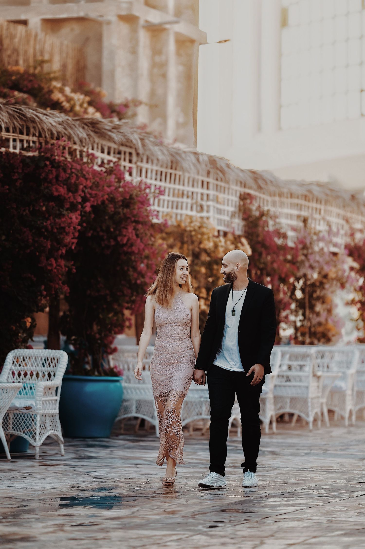 A couple strolls along a paved walkway with pink flowering bushes and white wicker furniture in the background.