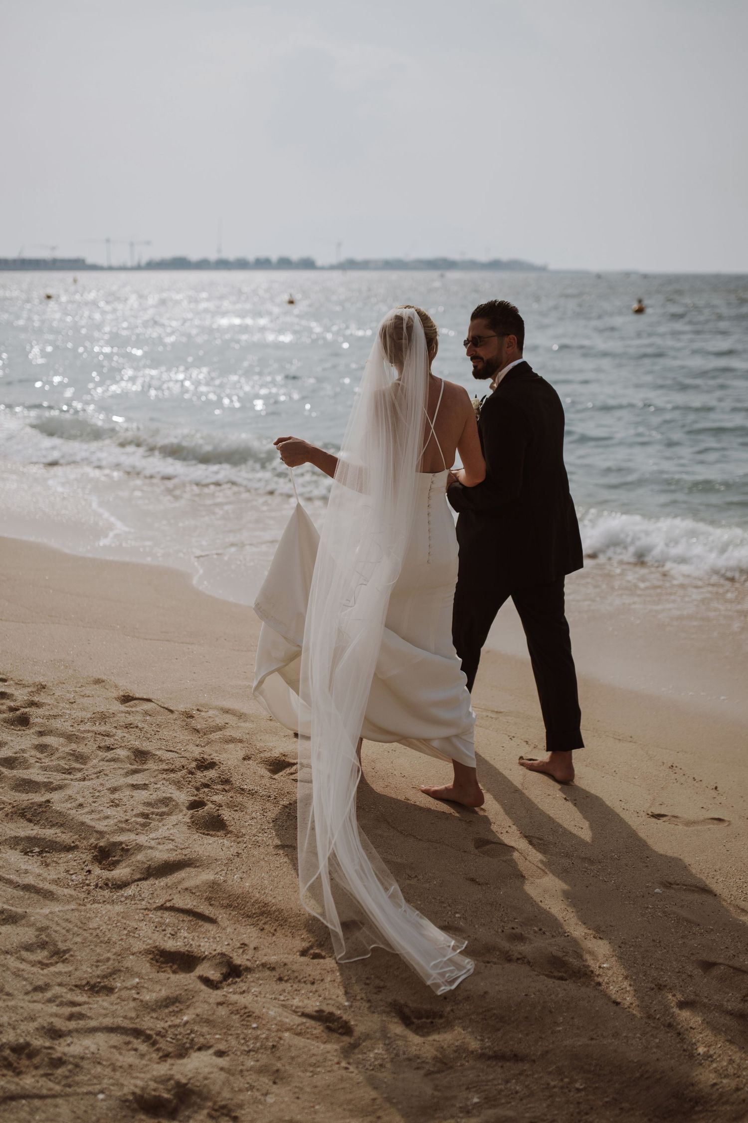 A couple in wedding attire walks along a beach shore with flowing veil and waves in the background.