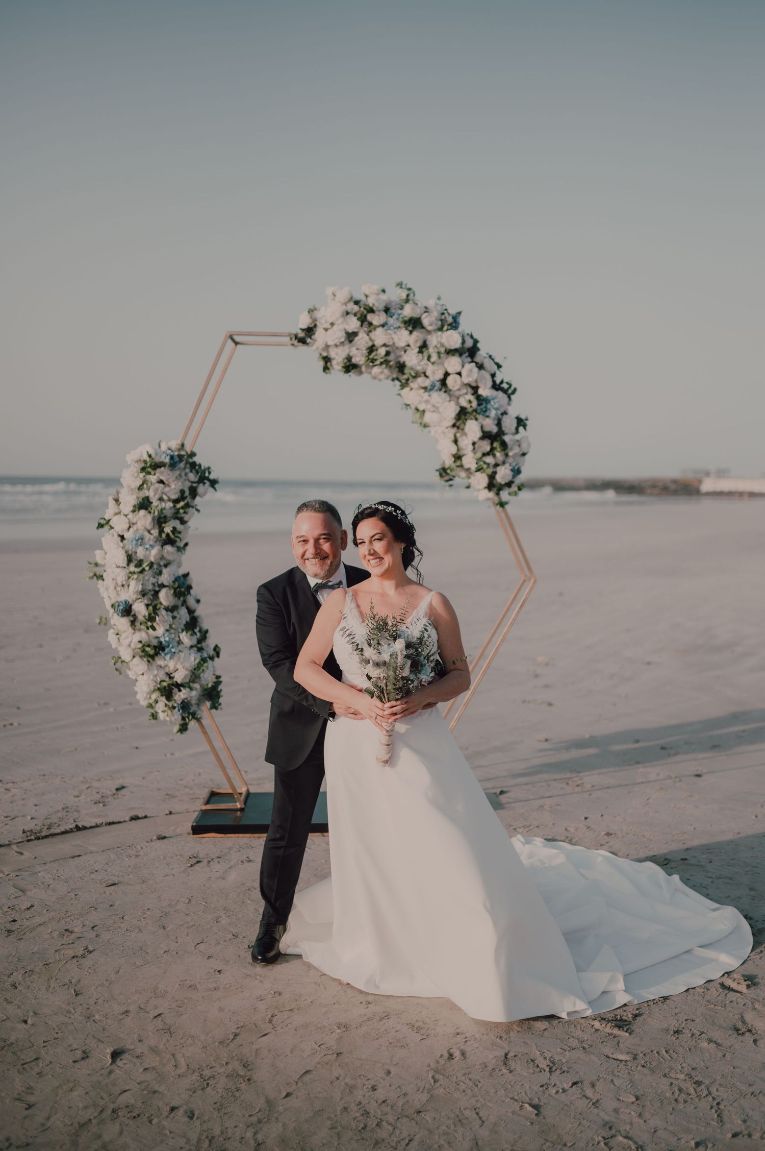 Beach wedding setup with floral arch and white decorations by the ocean.