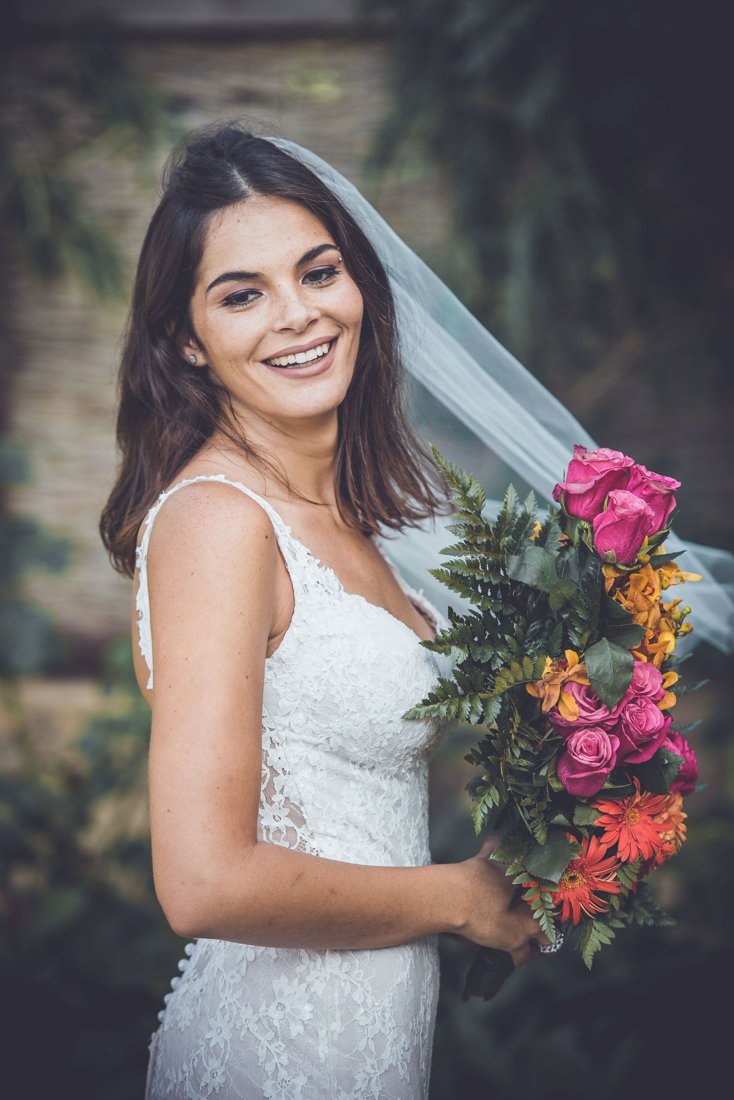 A bride in a lace wedding dress smiles while holding a vibrant bouquet of pink and orange flowers.
