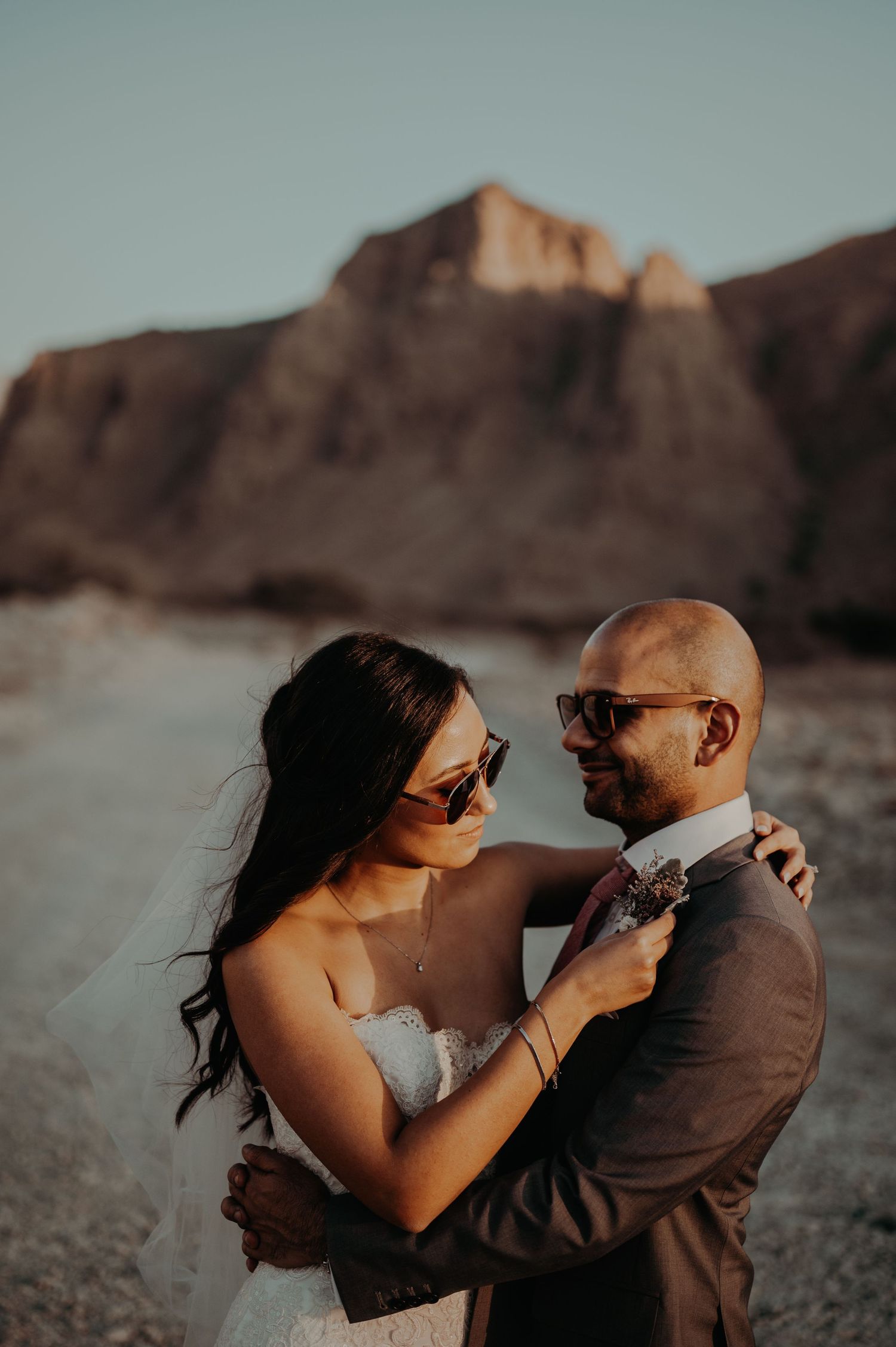 Couple in wedding attire embraces against backdrop of desert mountains at sunset.