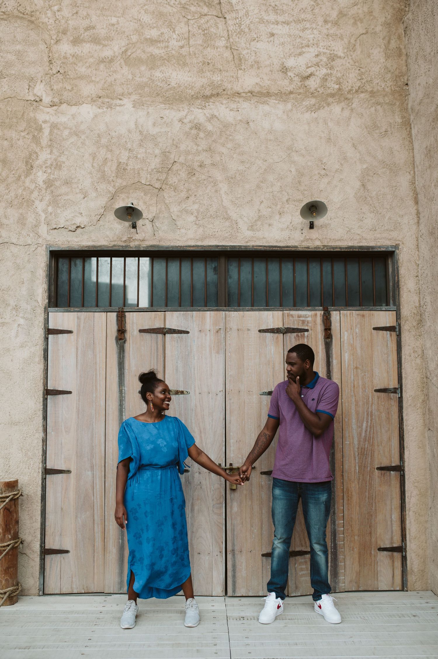 Couple in casual outfits posing playfully against a textured stone wall with wooden doors.