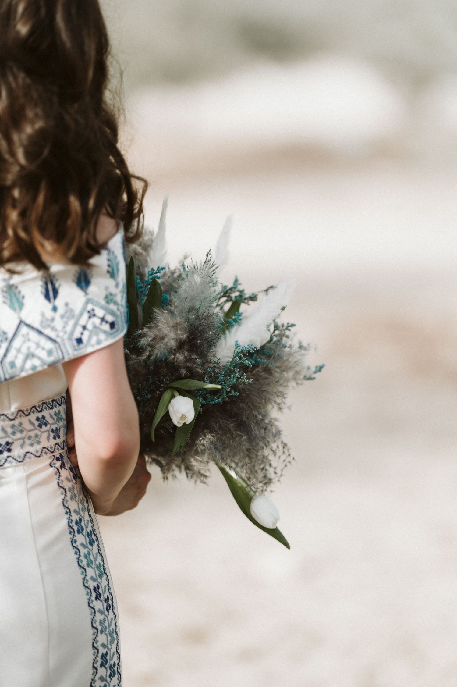A wedding bouquet with blue-tinted foliage and white flowers is held against a white dress with blue embroidery.