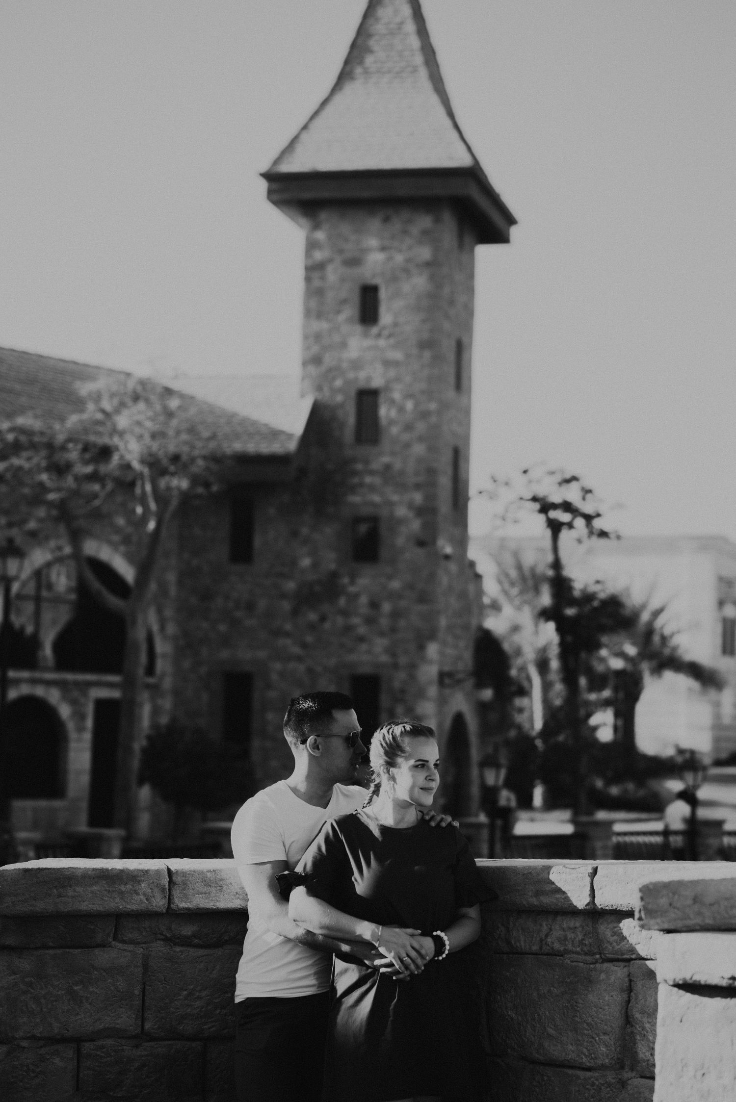 Couple sits together on stone wall near historic tower buildings in black and white photo.