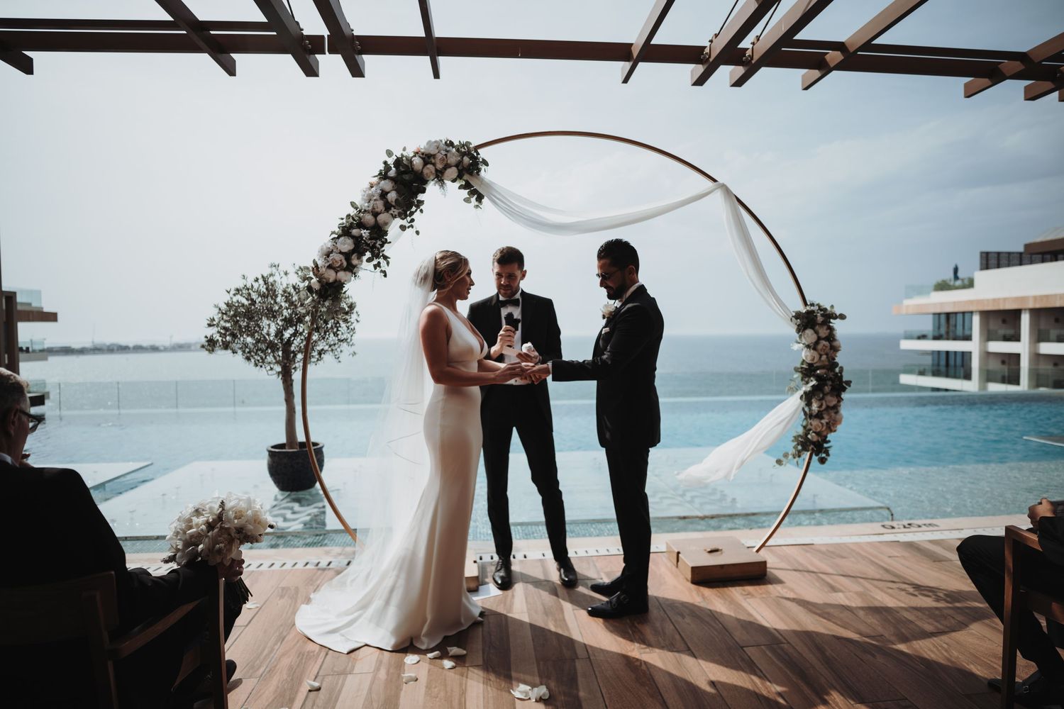 A wedding ceremony takes place under a wooden pergola overlooking the ocean.