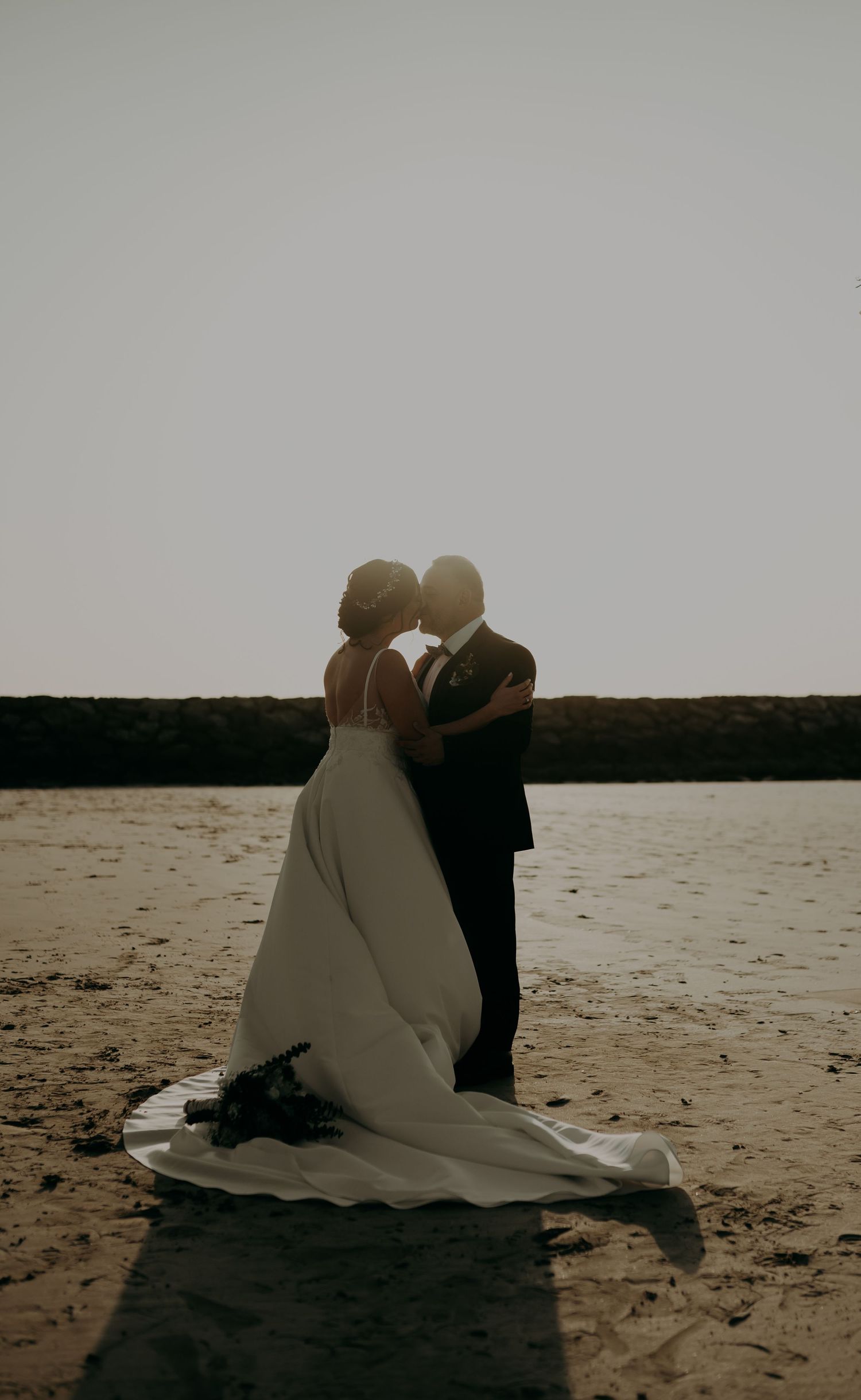 Silhouetted couple sitting on beach at sunset during wedding photoshoot.