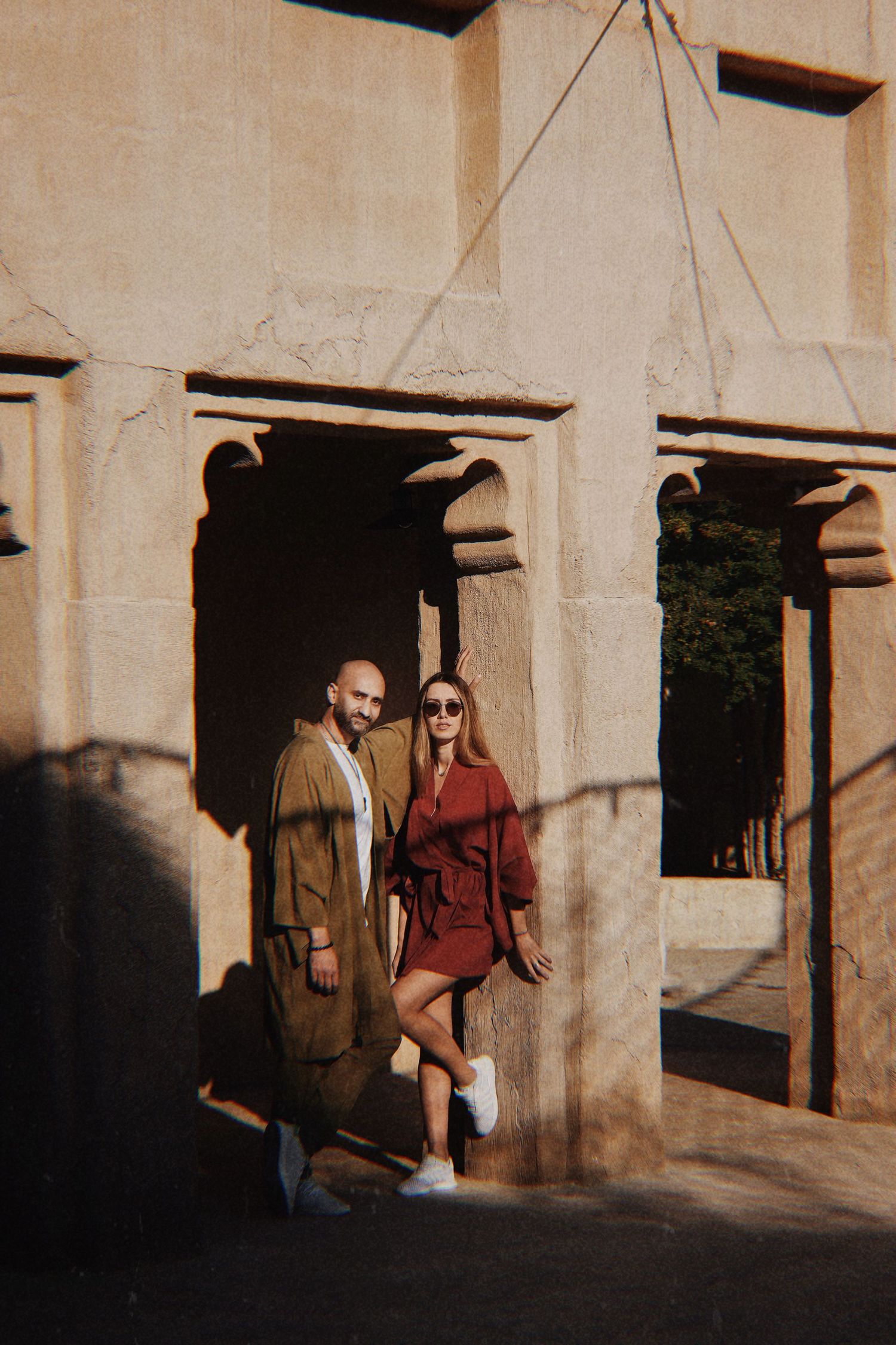Two people walk through a sunlit stone archway corridor casting dramatic shadows on the wall.