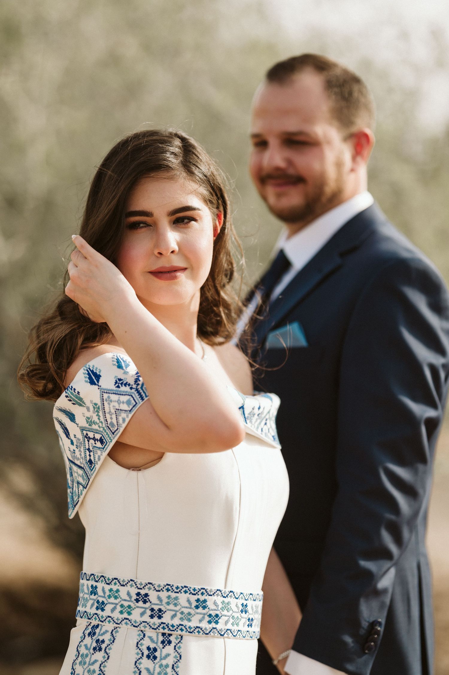A bride in a white dress with blue embroidery poses with her groom in a black suit outdoors.