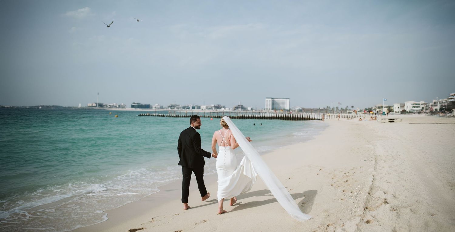 A couple walking on a beach with turquoise waters and cityscape in the background.