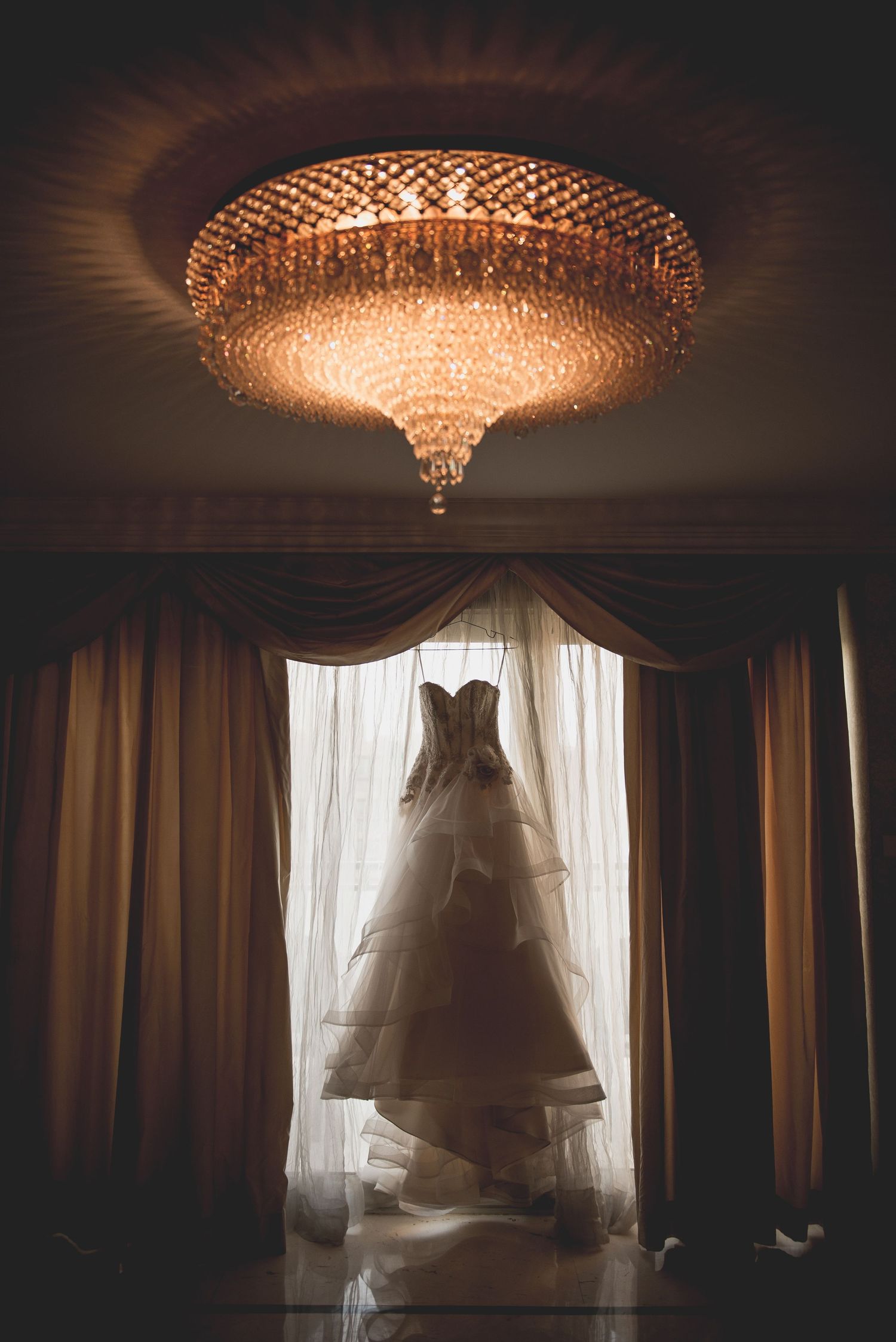 Wedding dress silhouetted against window with ornate crystal chandeliers in luxury hotel room.