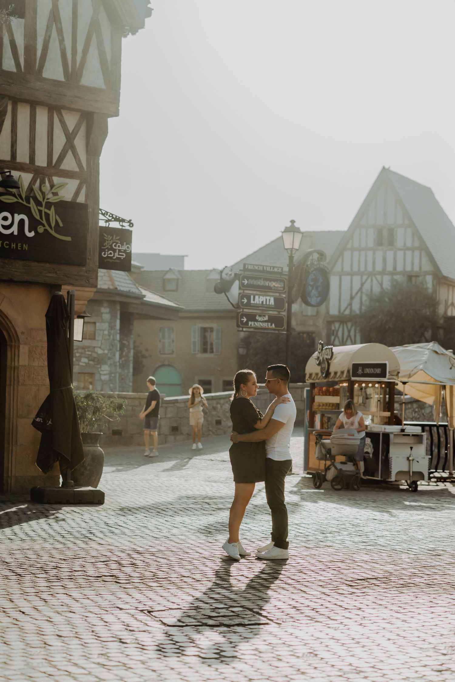 Two people dance together on a cobblestone street in front of vintage Tudor-style buildings on a misty day.
