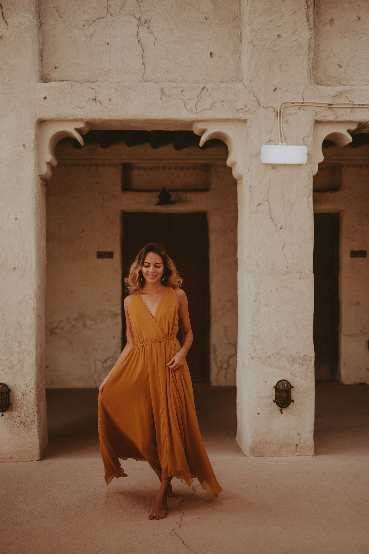 A golden dress twirls gracefully through a series of white stone archways in warm lighting.