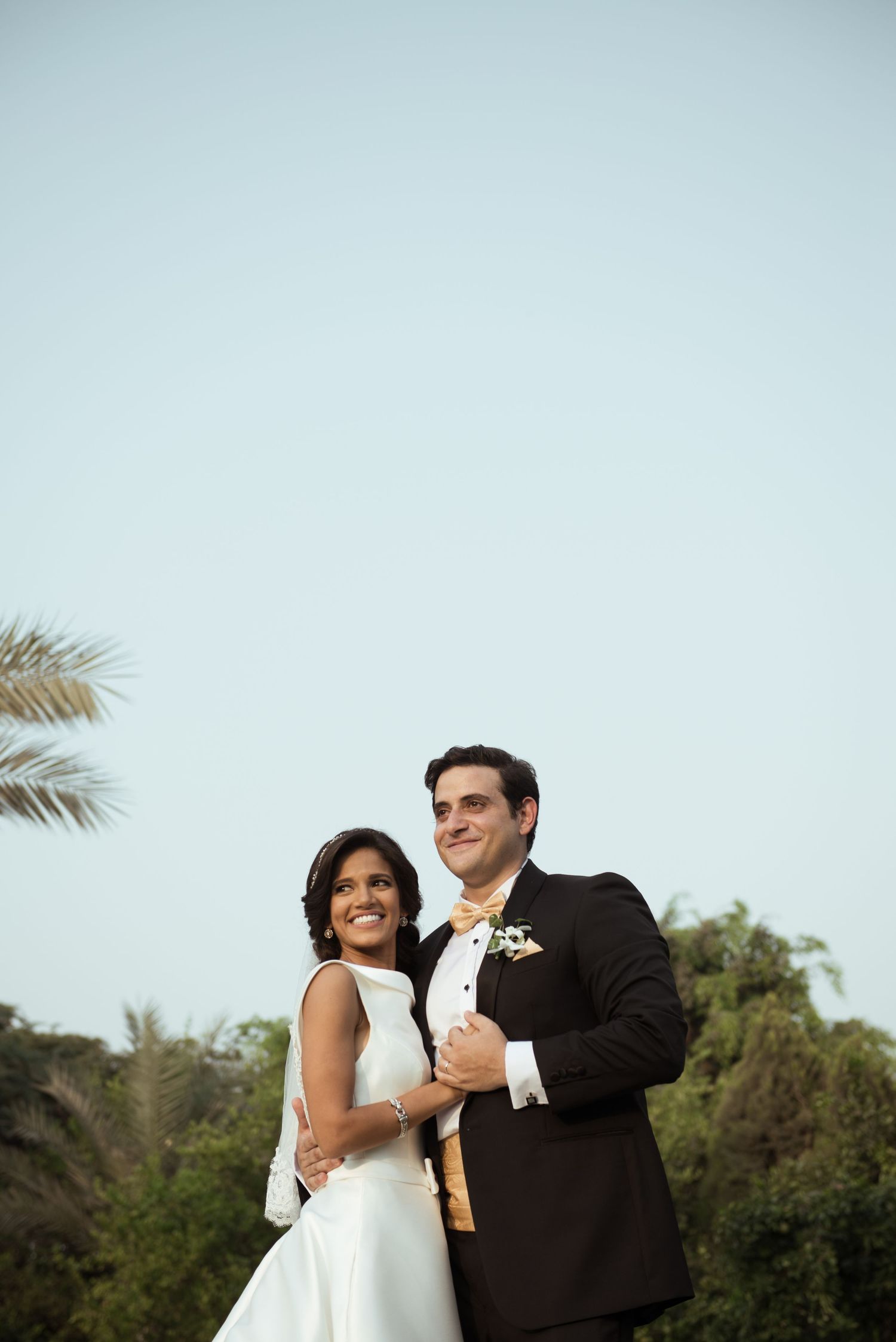 Elegant wedding couple poses against a light blue sky with palm trees in the background.