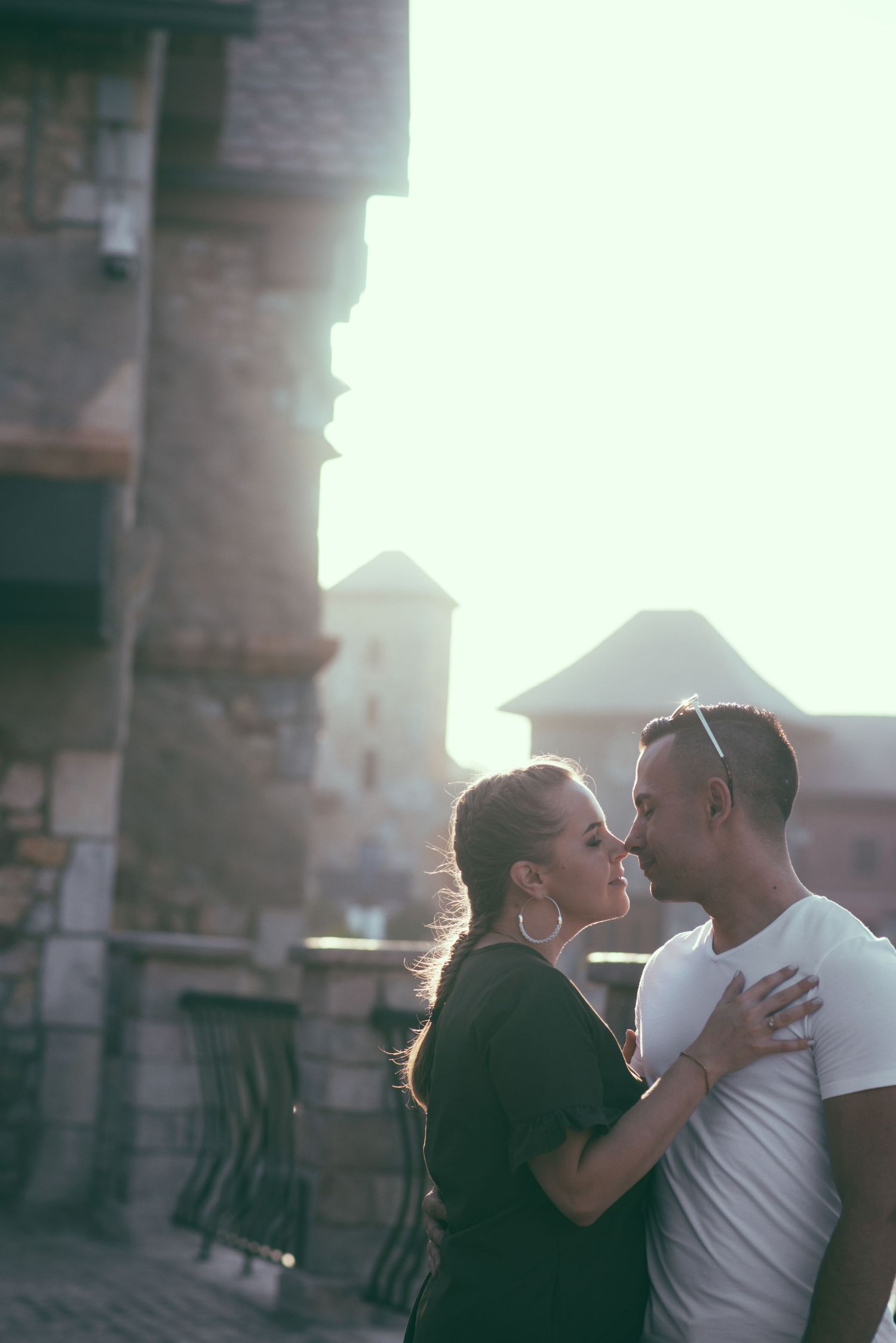 A couple embraces in a romantic moment against a sunlit urban backdrop with brick buildings visible.