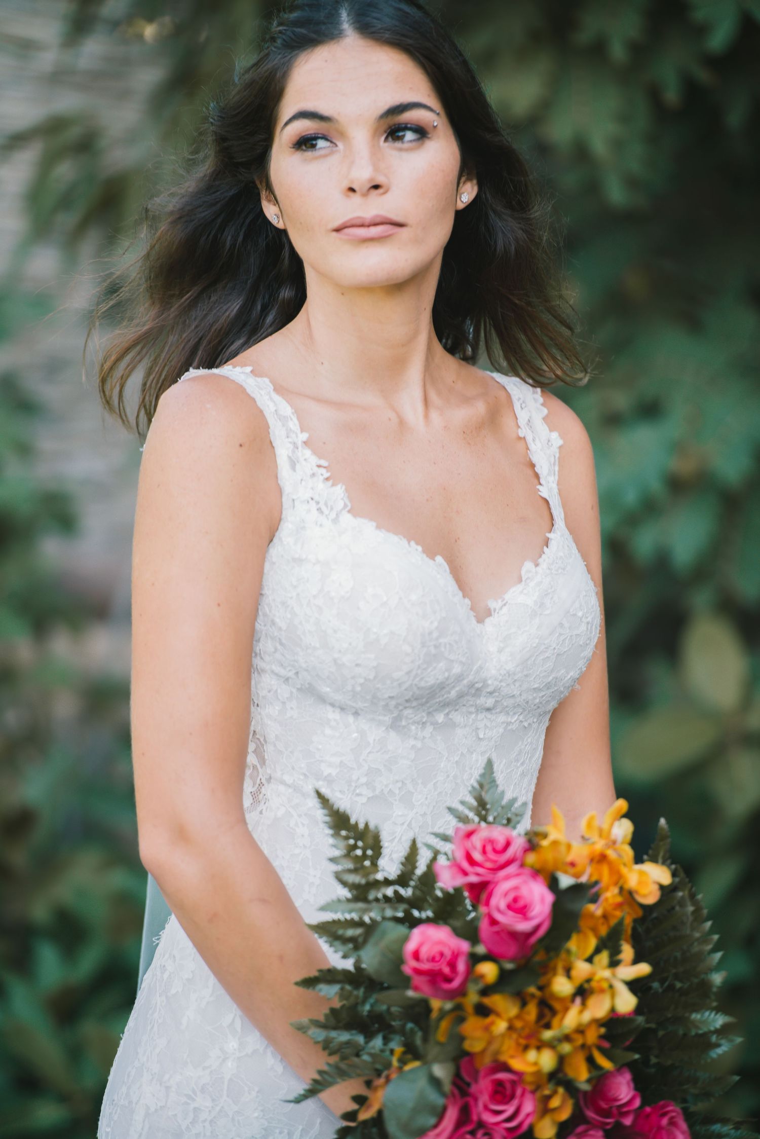 A bride in a white lace dress poses with a colorful bouquet against a garden backdrop.