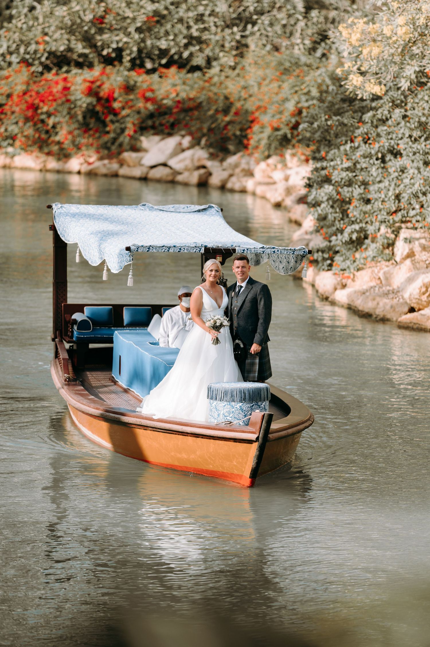 Newlyweds ride in a covered wooden boat along a scenic waterway with flowering bushes.
