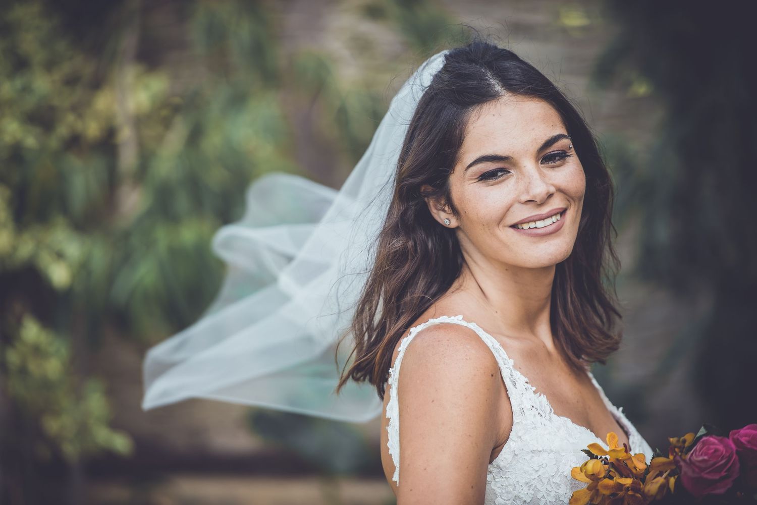 A bride in a white lace dress holding a colorful bouquet with her veil flowing in the wind.