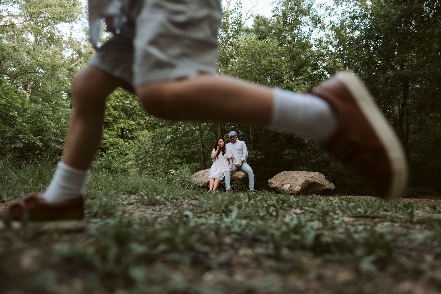 A couple sits together on a blanket in a serene outdoor setting surrounded by lush greenery and natural light.