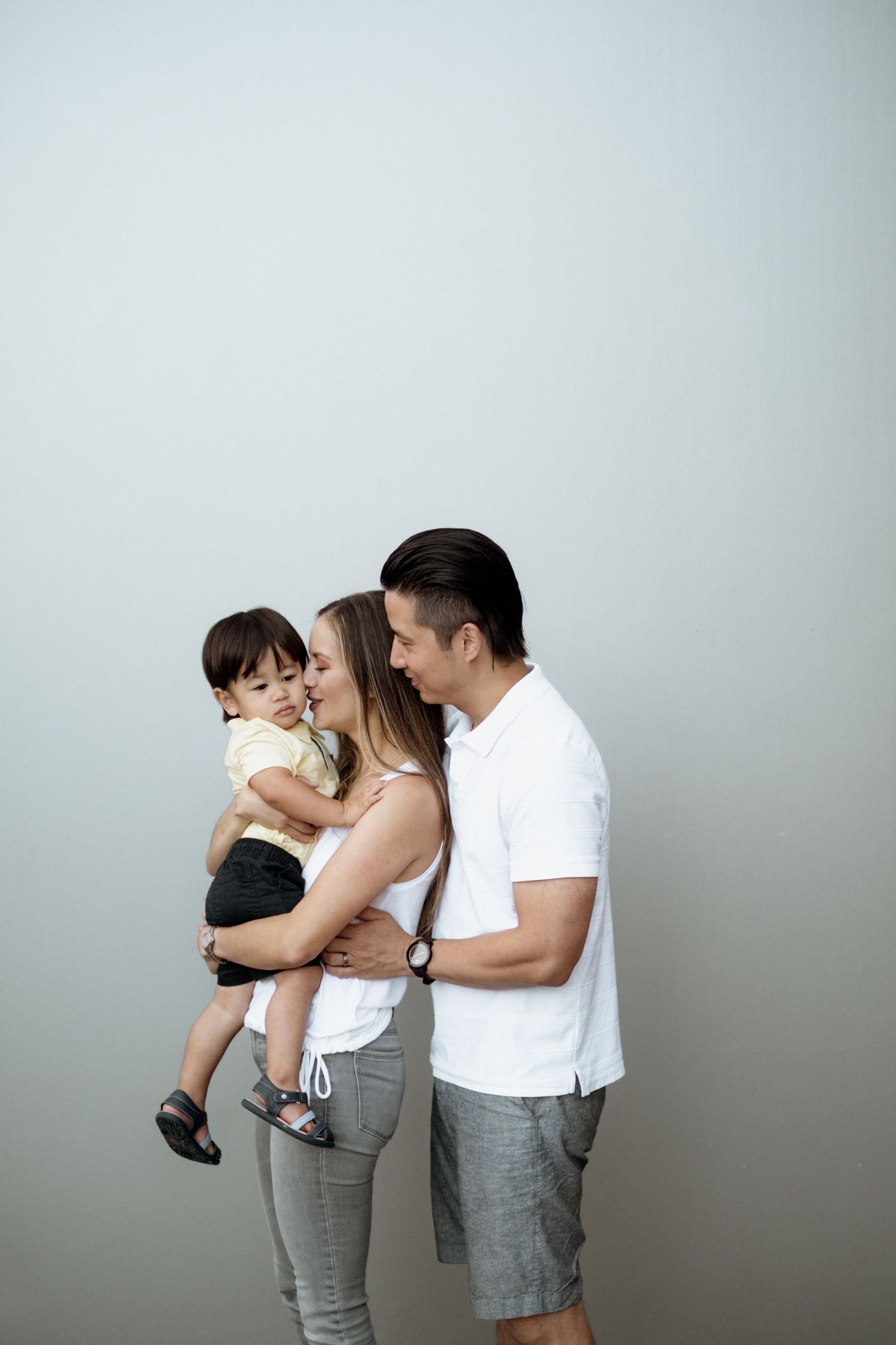Family in white and gray casual wear share tender moments in minimalist studio setting against neutral background.
