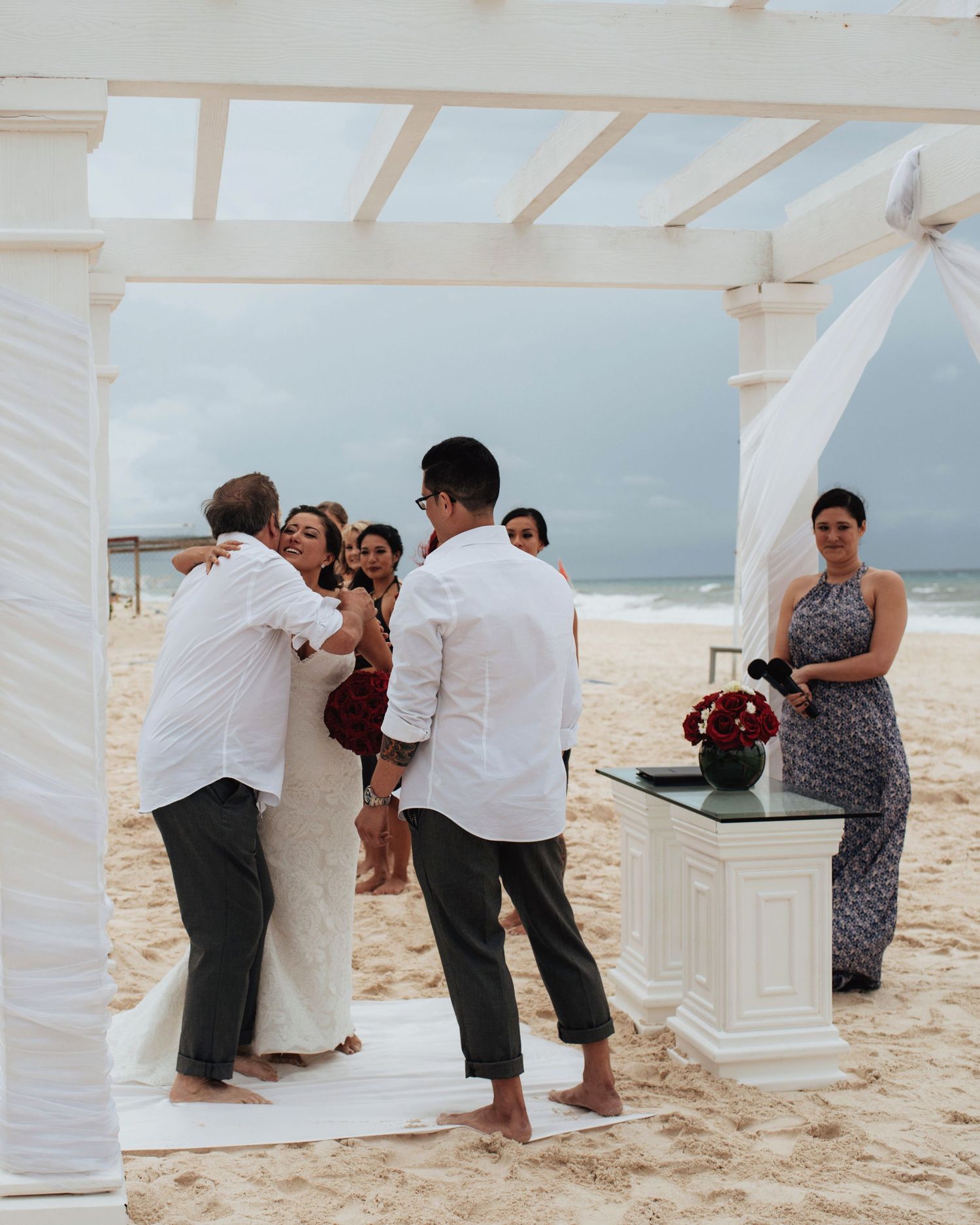 Wedding guests gathering around a white pergola structure on a sunny beach.