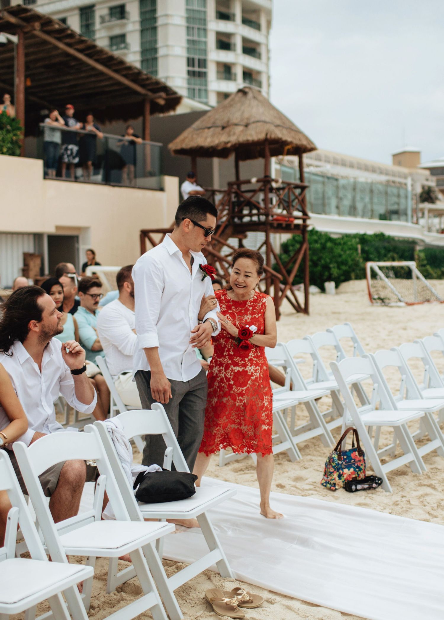 Beach wedding setup with white chairs and a thatched umbrella against oceanfront buildings.