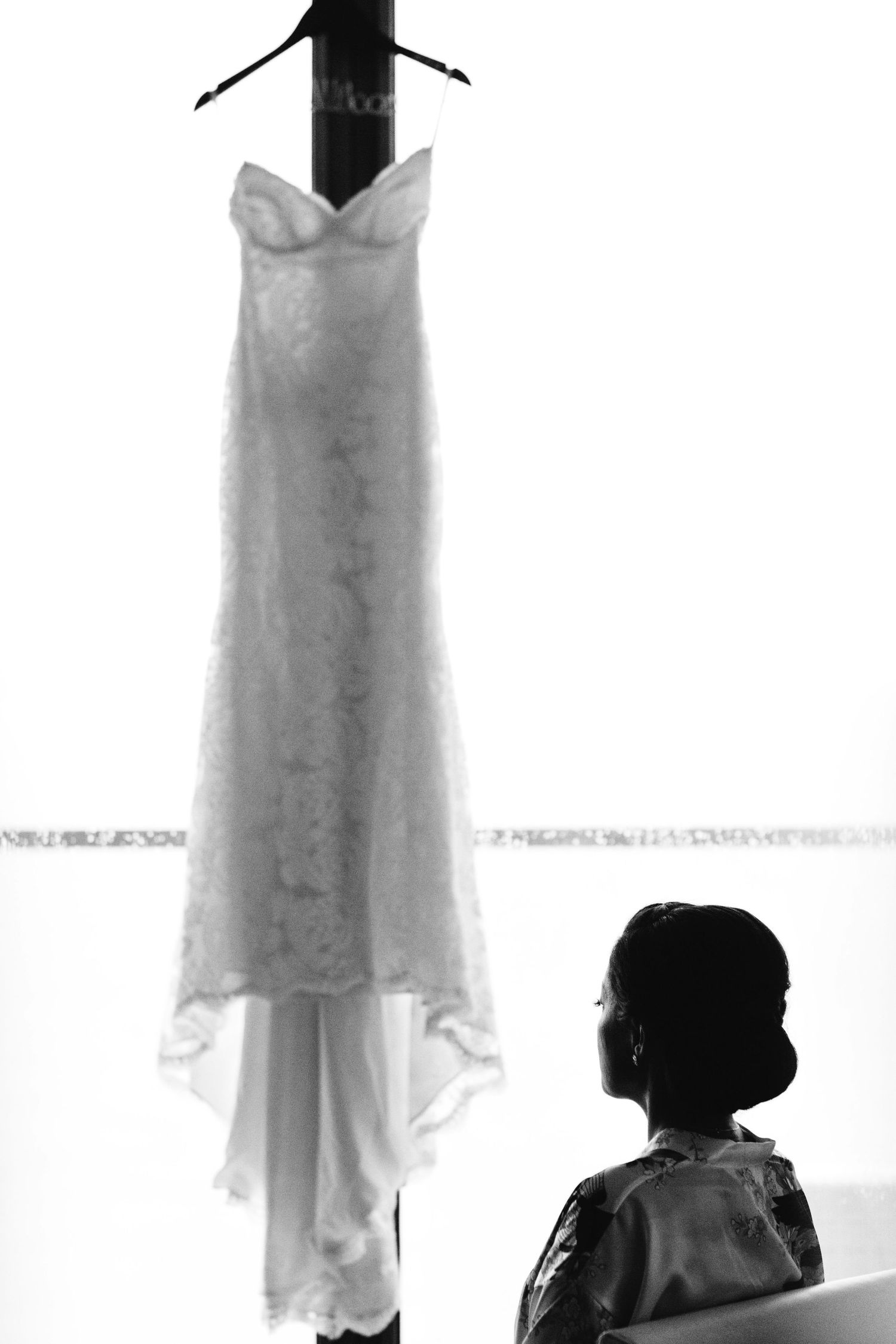 A white wedding dress hangs elegantly against a bright window backdrop.