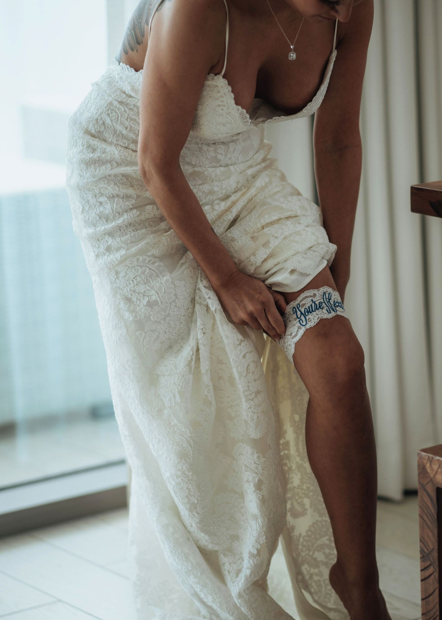 Person in wedding dress attaching a garter to their leg near a window.