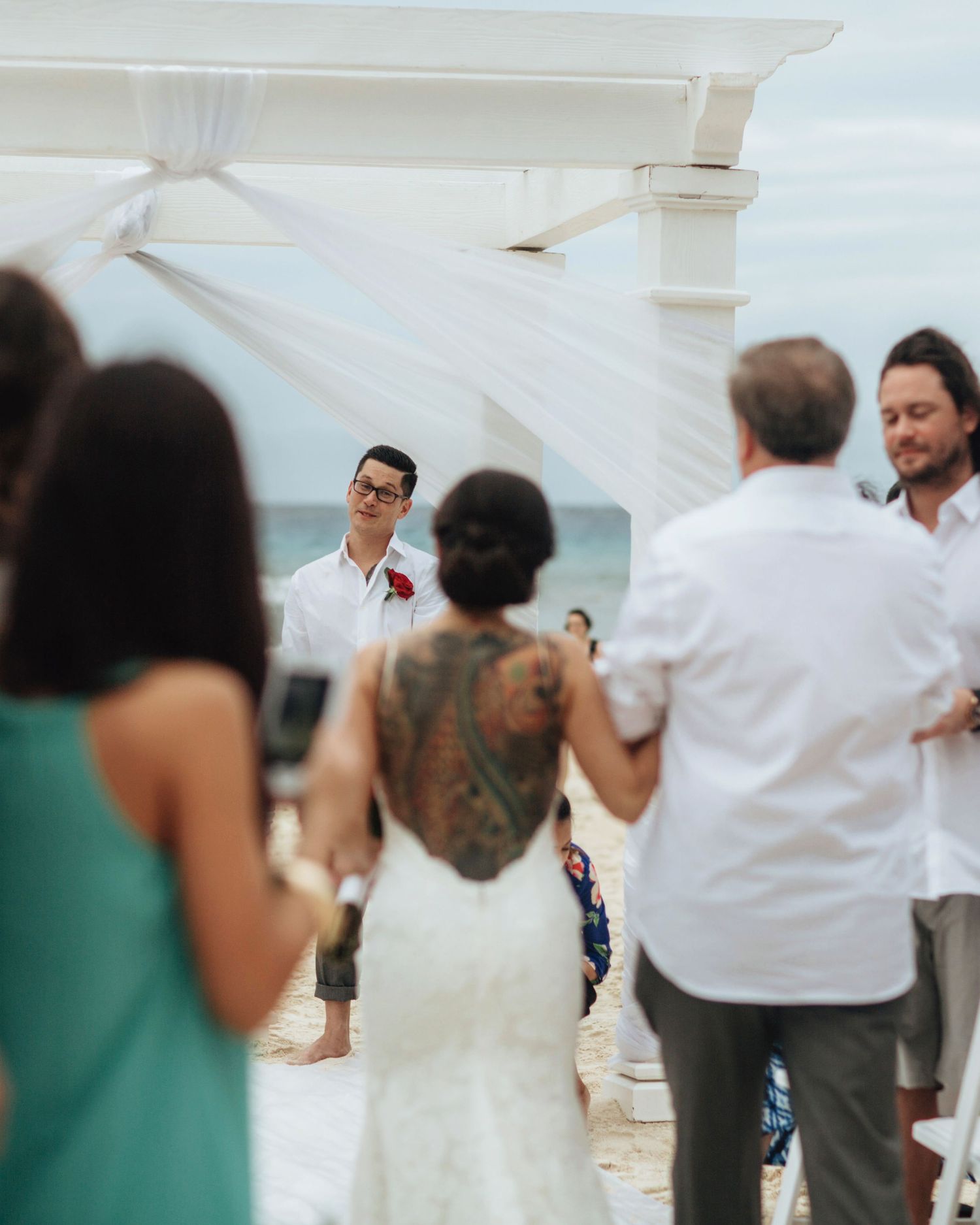 Wedding ceremony taking place under a white pergola on the beach with guests watching.
