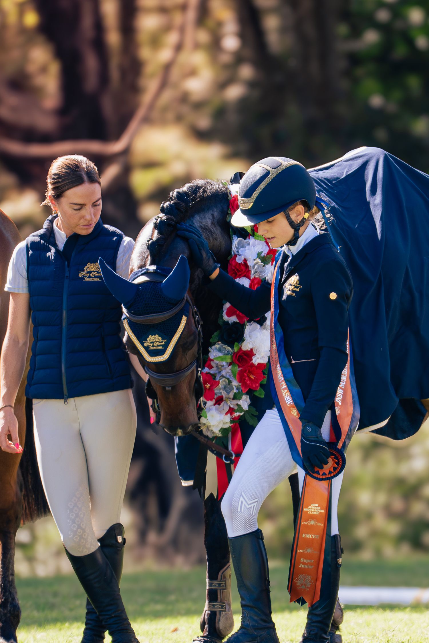 Equestrian riders in navy jackets and white pants stand together with their horse adorned with red and white flowers.