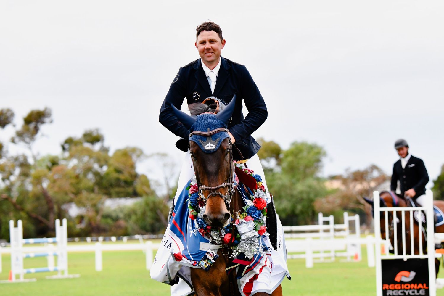 Rider smiling and cantering towards the camera with the horse wearing decorative floral garlands at an equestrian competition.