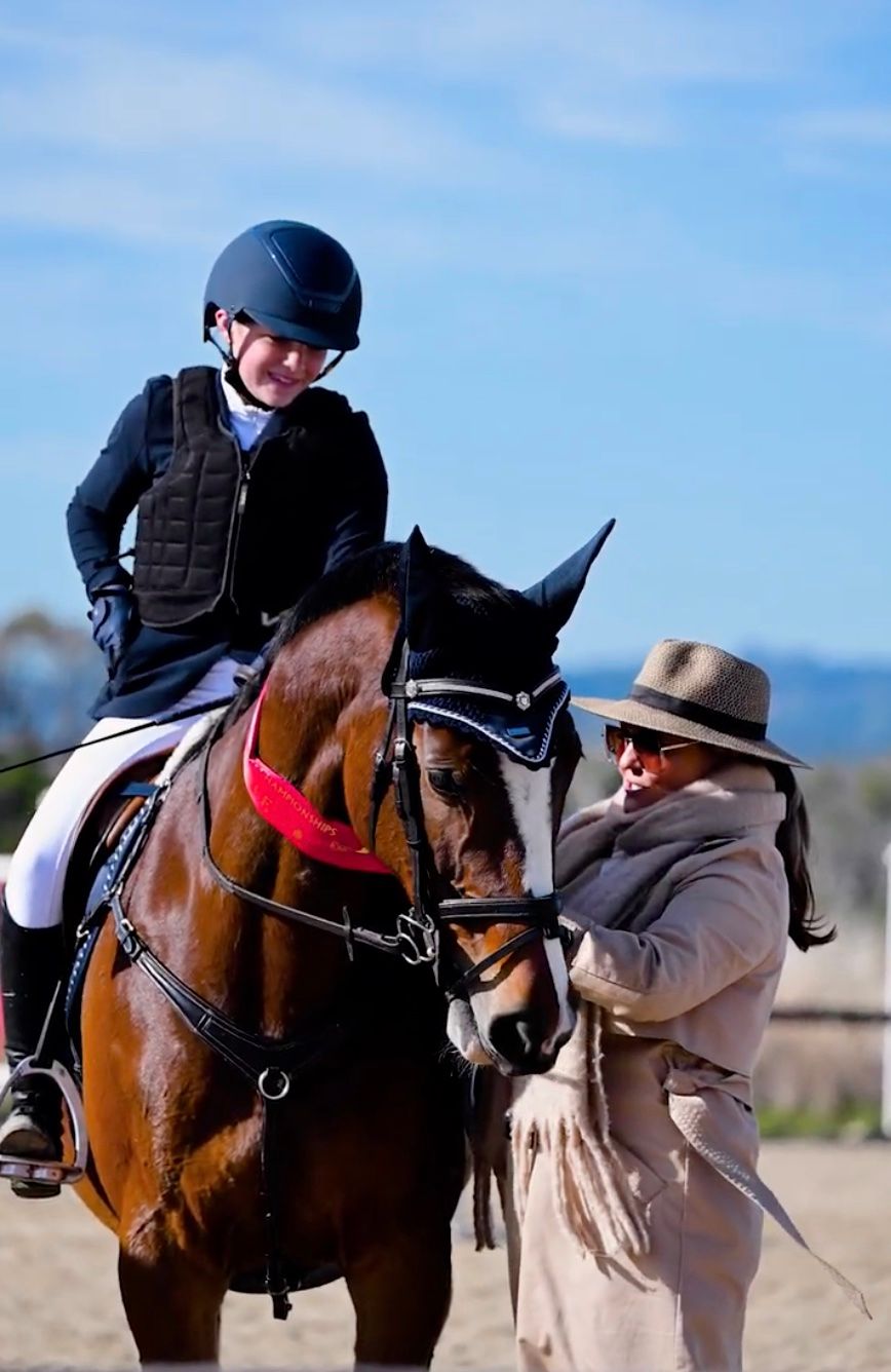 Show jumping judge tieing red ribbon onto bay horse's neck with rider in saddle, smiling.