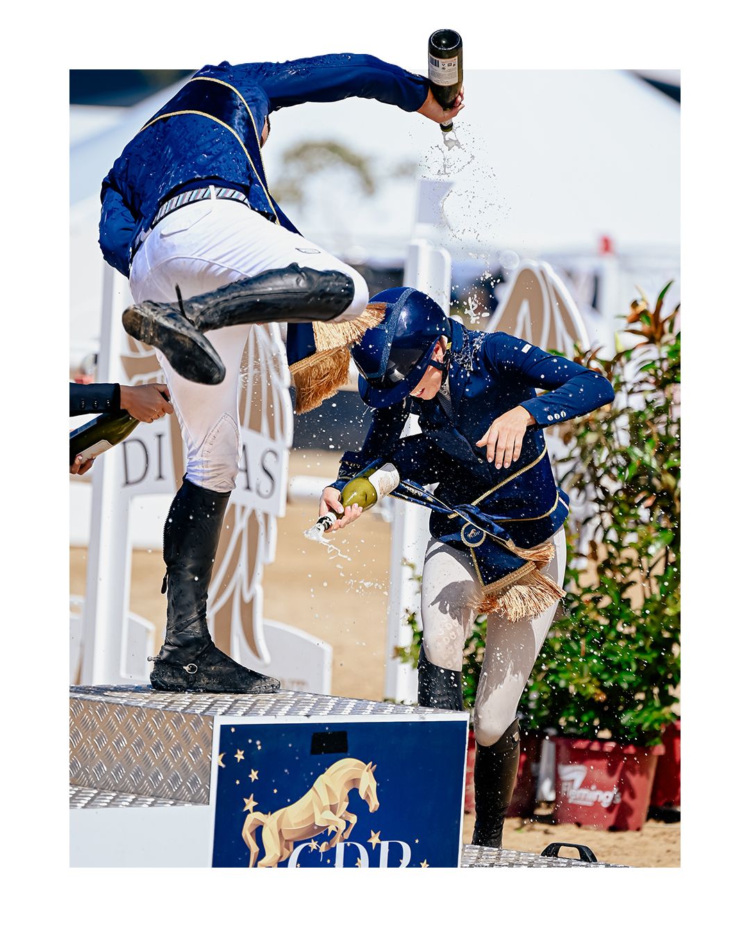 Two equestrians in blue uniforms celebrate with champagne spray at a show jumping podium ceremony outdoors.
