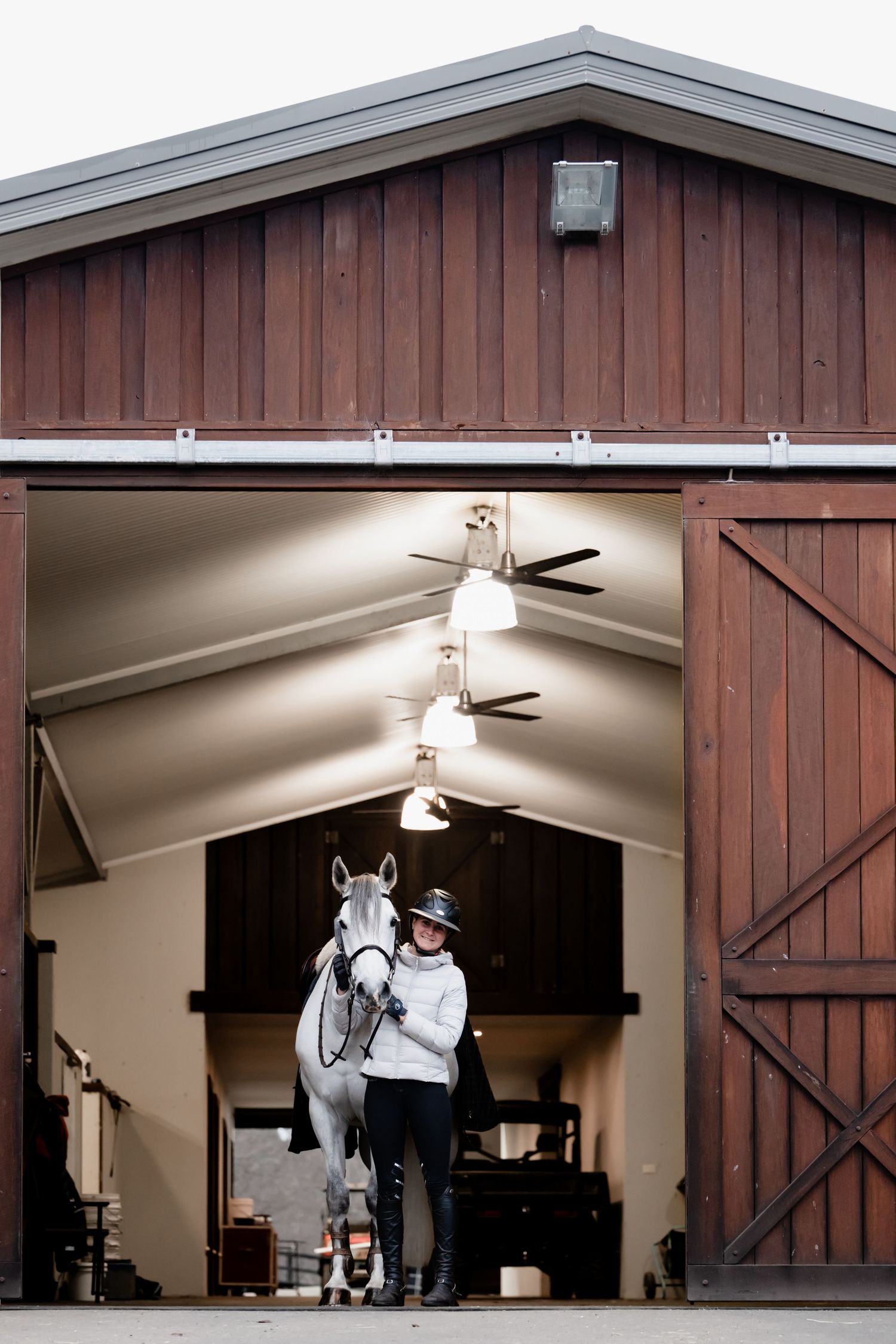 A white horse stands with its handler in the entrance of a rustic brown barn with sliding doors and ceiling fans.