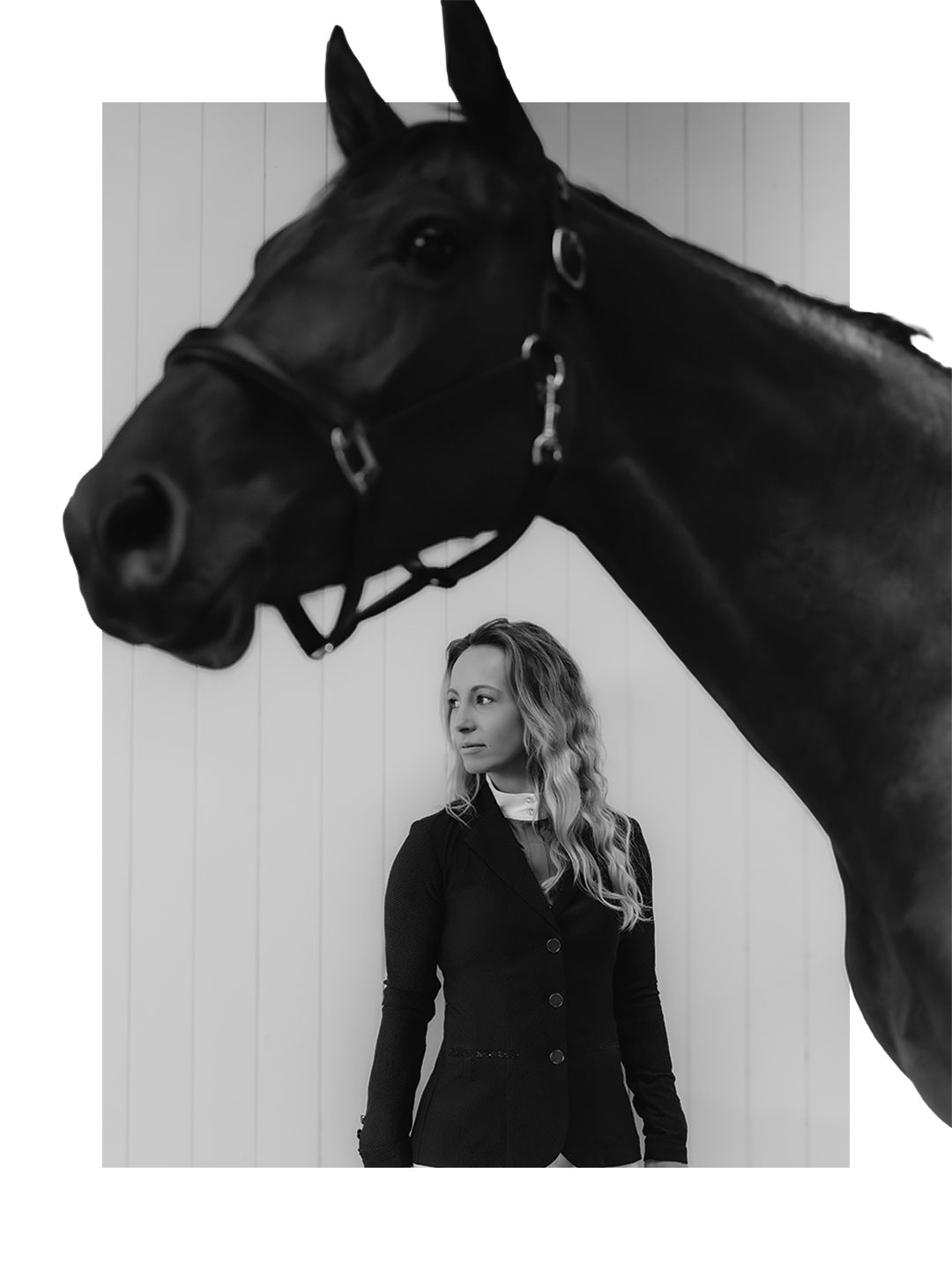 Black and white portrait of a blonde woman in equestrian attire standing beside a dark horse near a white wall.